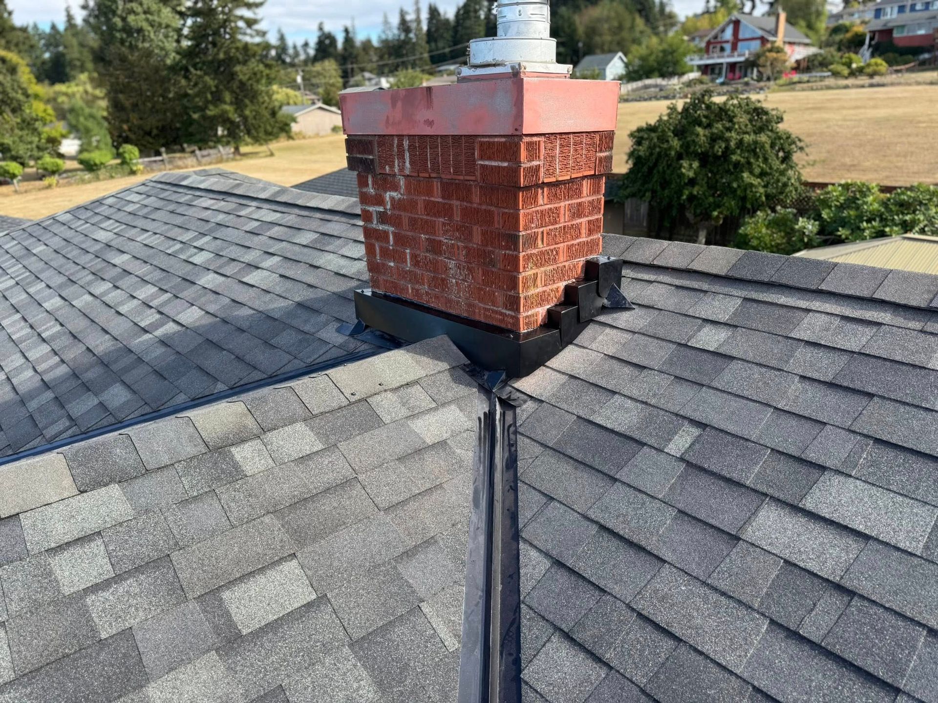 Brick chimney on a gray shingle roof, with flashing and a white chimney cap, set in a neighborhood.