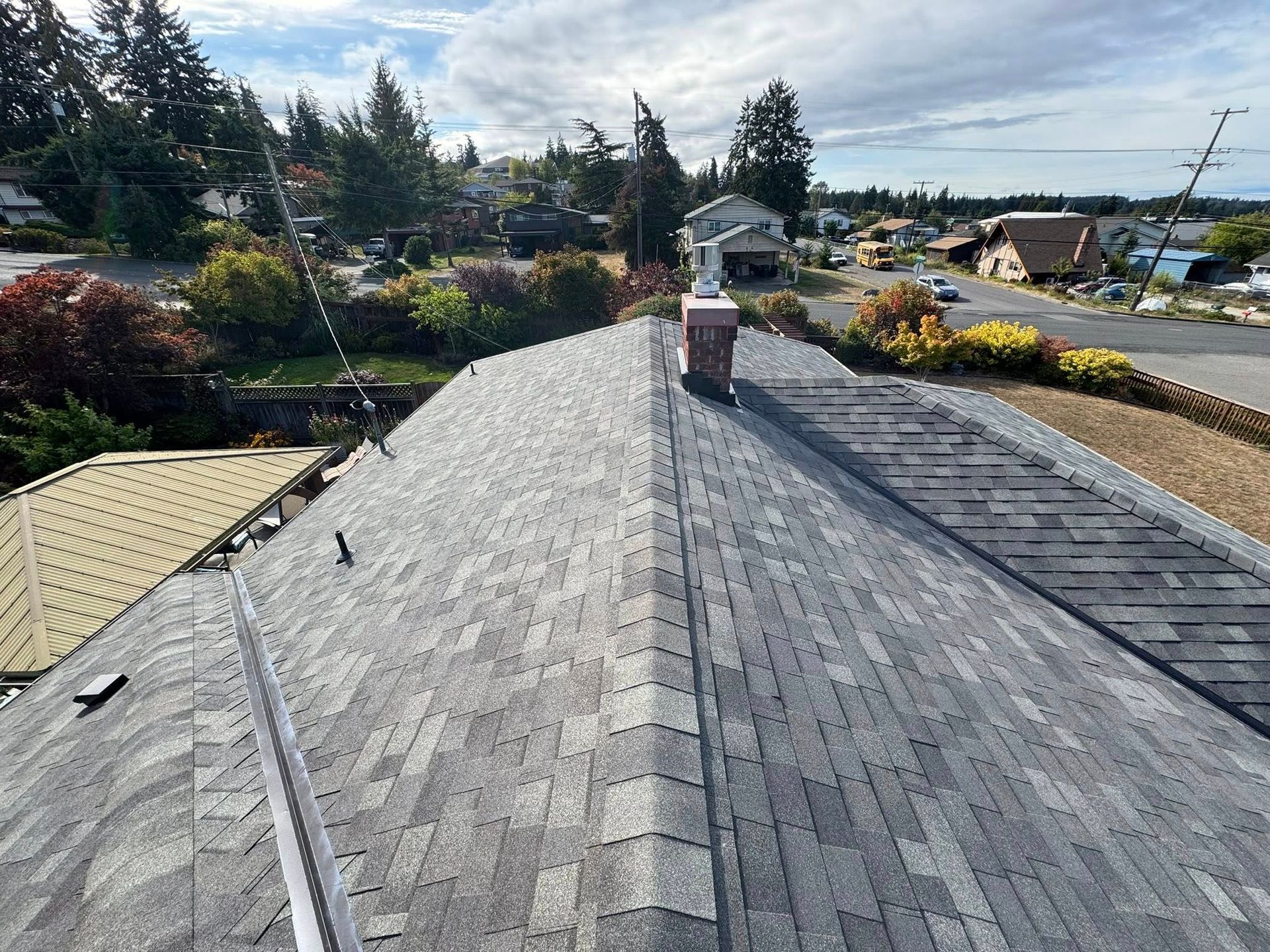 Overhead view of a gray shingle roof with a brick chimney and neighborhood in the background.