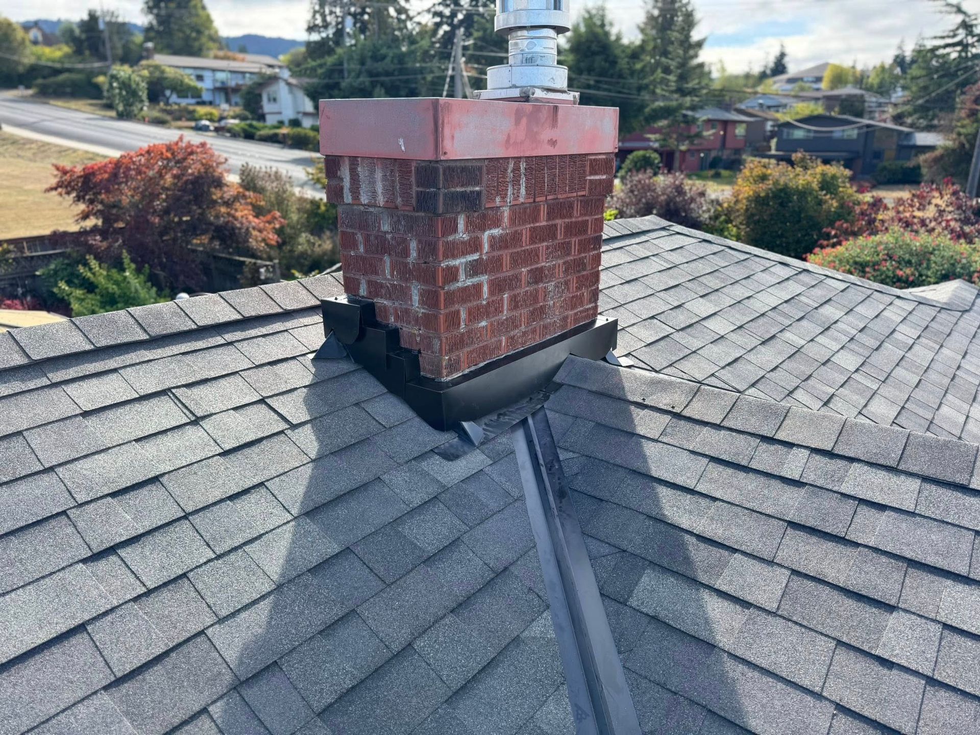 Brick chimney on a gray shingled roof, with visible metal flashing and a suburban backdrop.