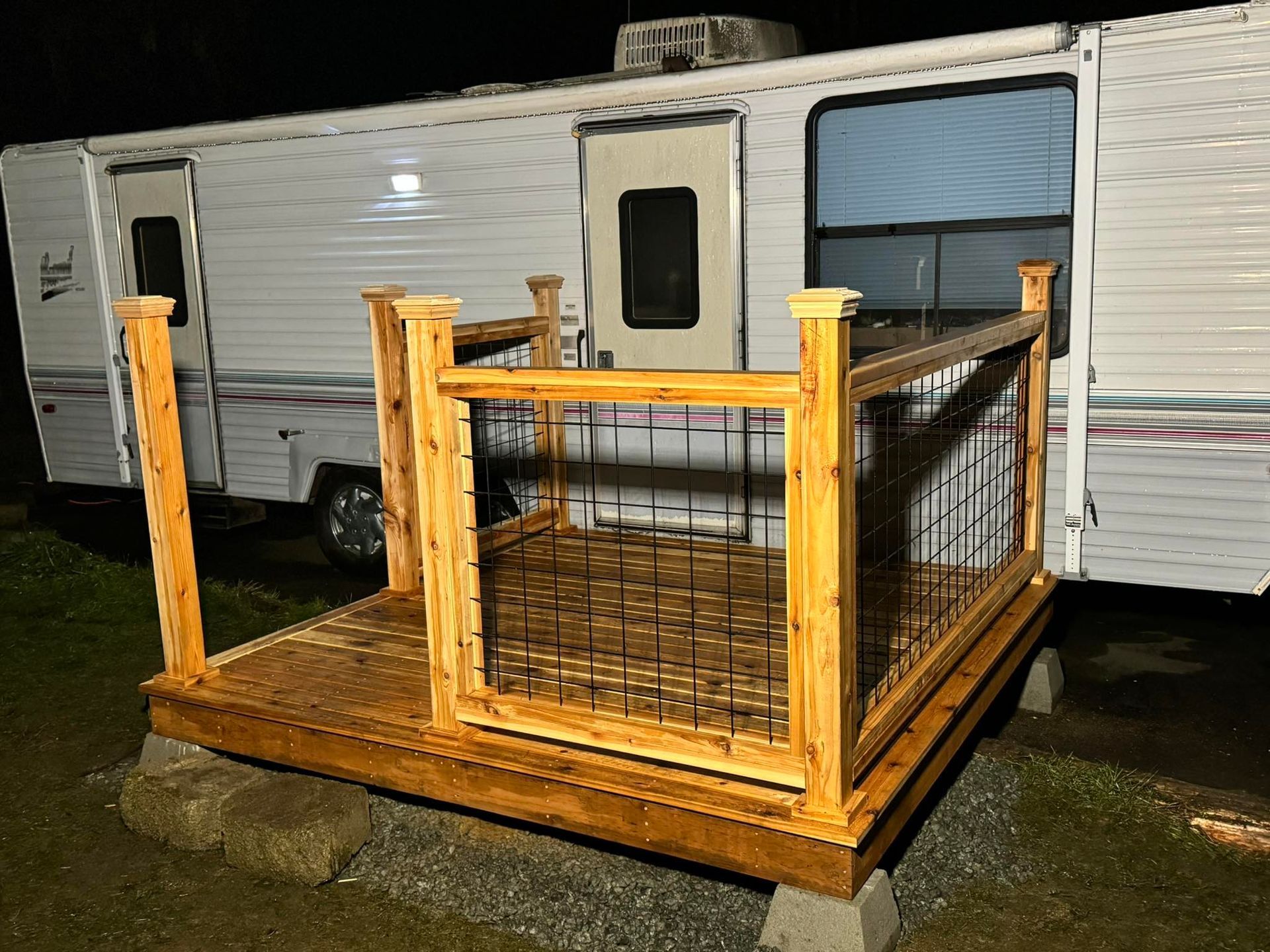 A wooden deck with railings attached to a white RV, on a cement block base.