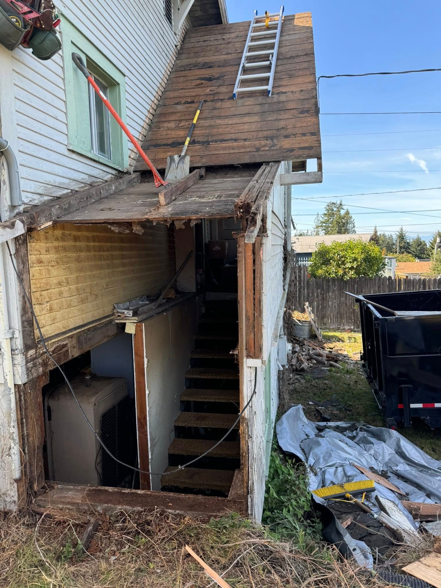 Exterior house renovation. Exposed staircase, damaged roof section, ladder, and a dumpster.