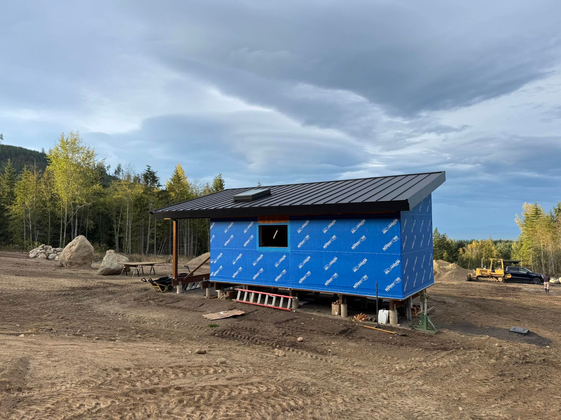 A small building under construction, wrapped in blue protective sheathing. Set on a dirt lot, cloudy sky above.