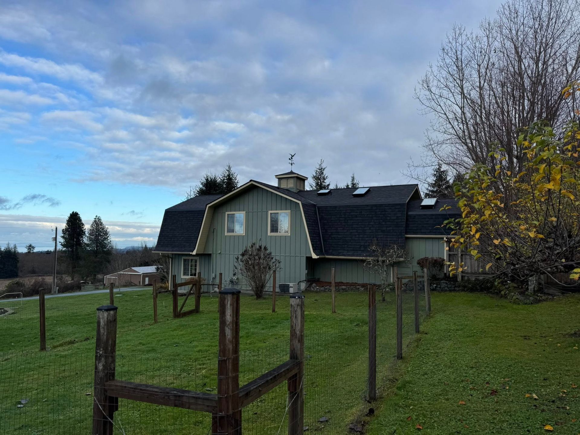 Green barn with black roof and small cupola, behind a wooden fence, under a cloudy sky.