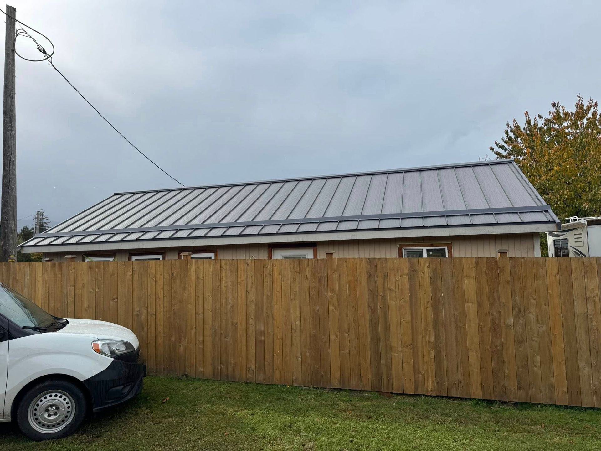 Building with metal roof behind a wooden fence, cloudy sky. White vehicle parked in front.