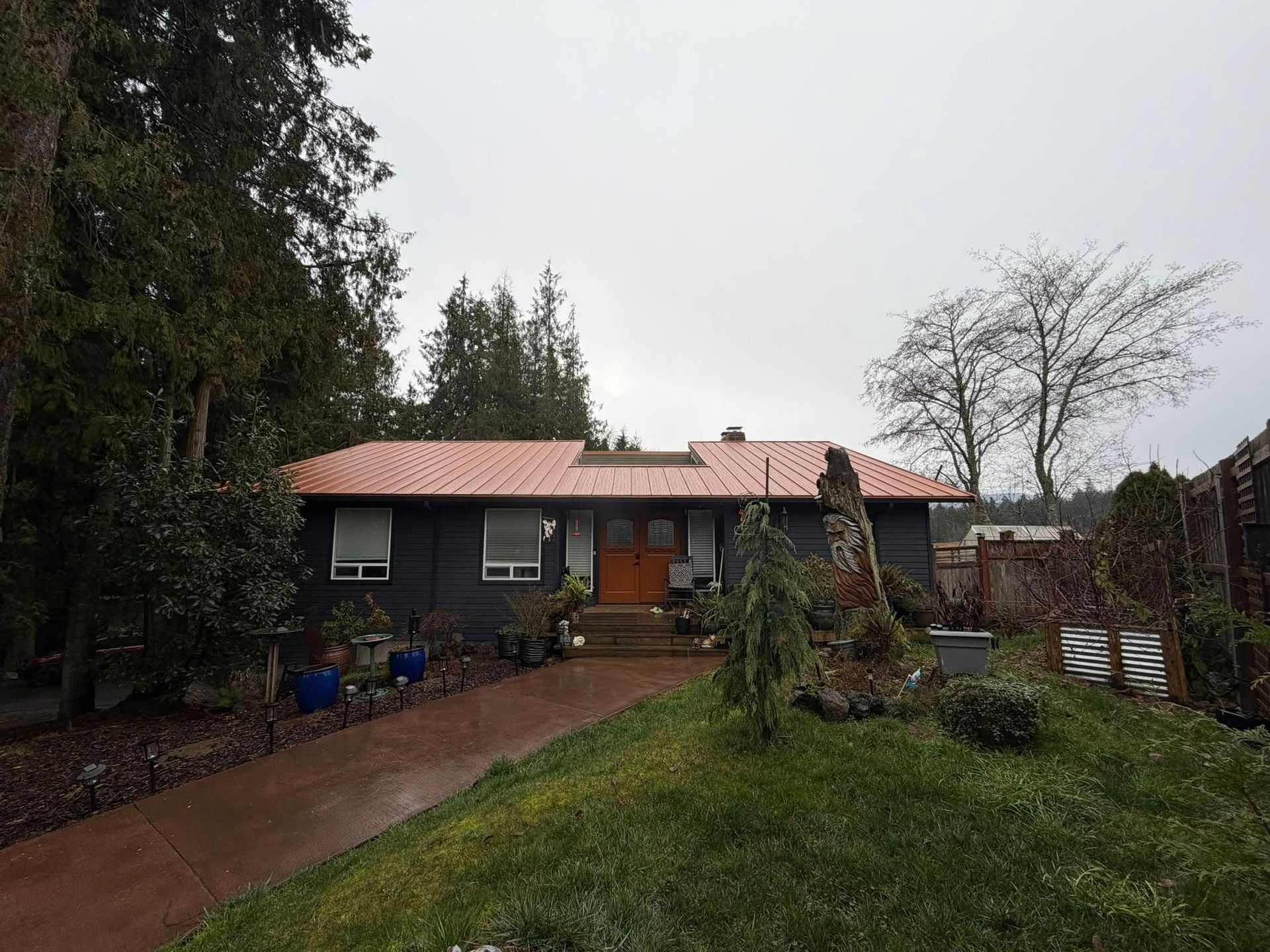 Gray house with orange roof, brown driveway, and green lawn. Tall trees surround the house.