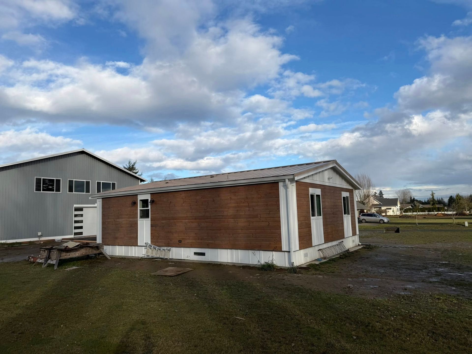 Mobile home with brown siding, white trim, and a gray roof sits on a grassy lot under a cloudy sky.