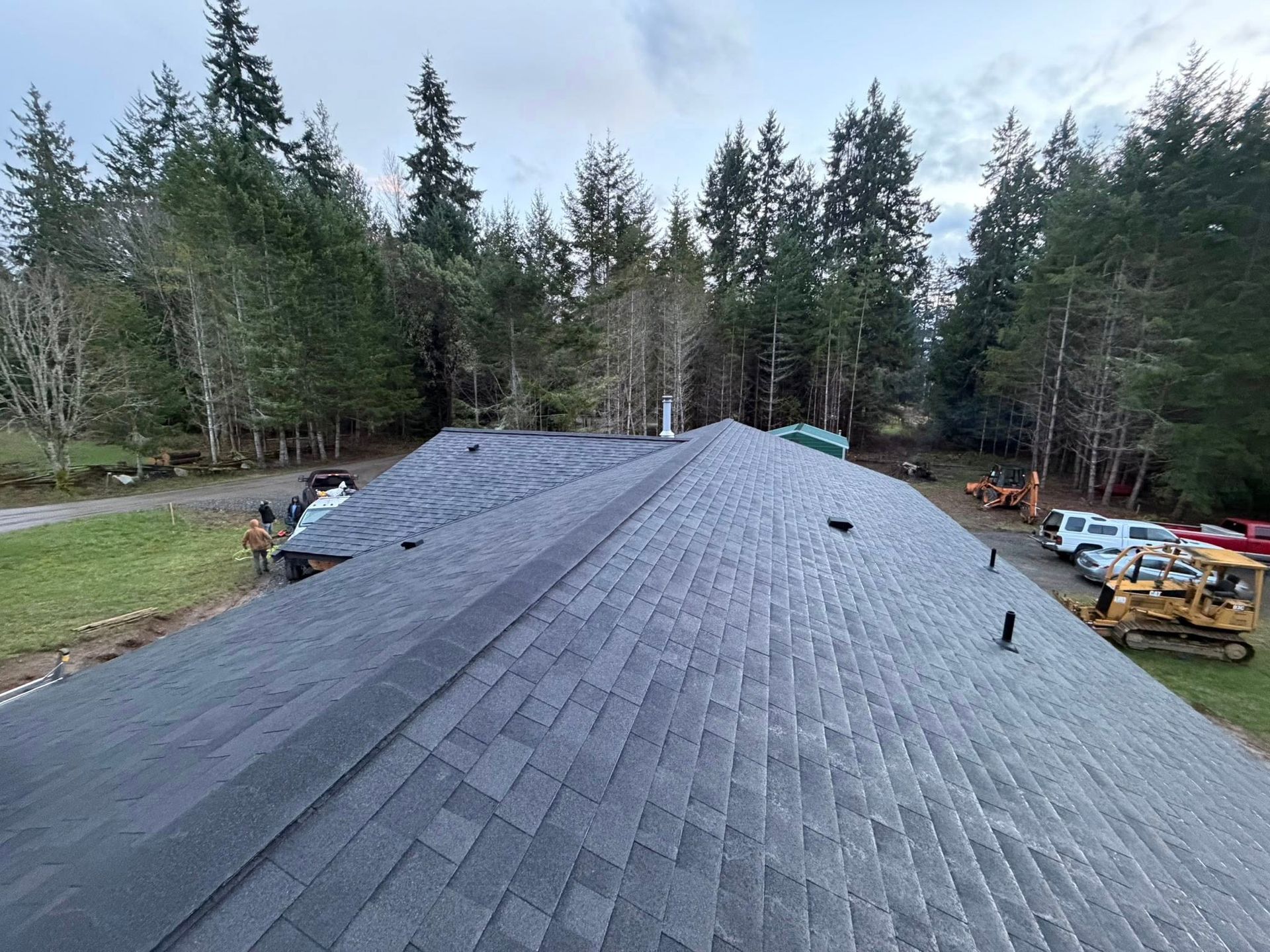 Newly shingled, gray roof on a house, forest background, construction equipment nearby.