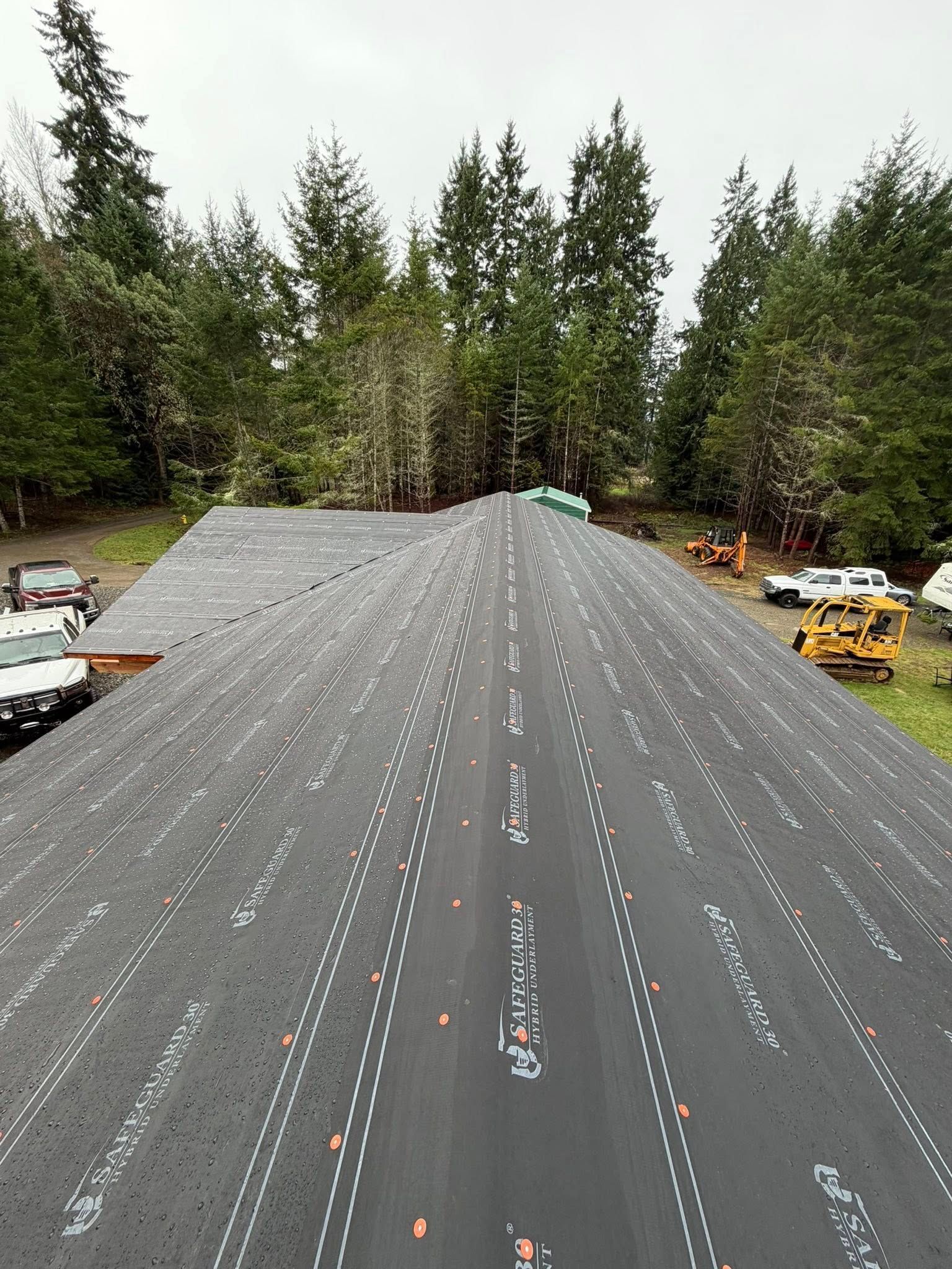 A partially shingled roof with trees in the background, construction equipment nearby.