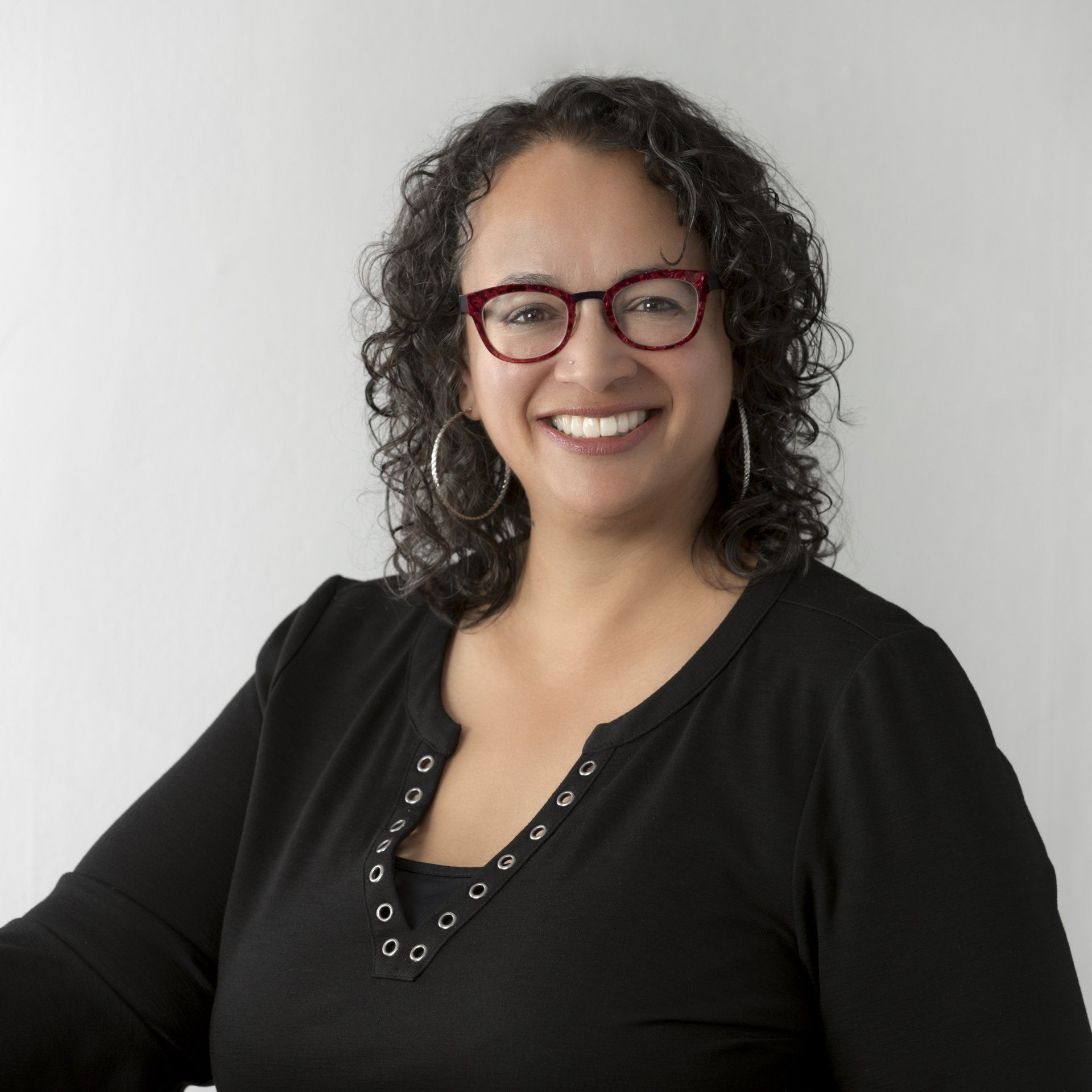 Smiling woman with curly hair and red glasses wearing a black top against a white background