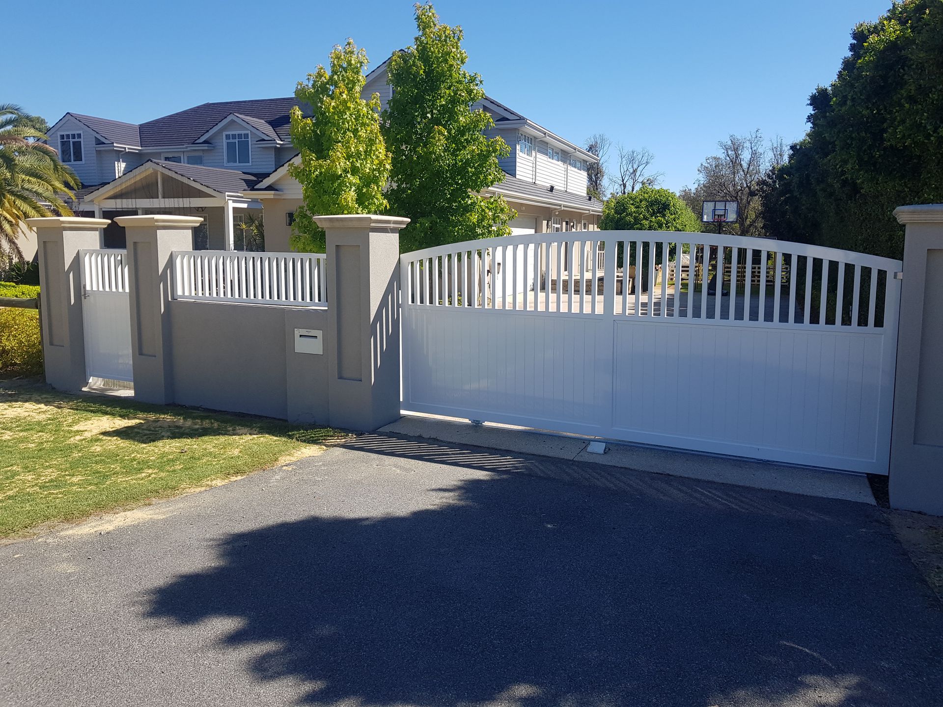White sliding gate in front of a gray house, with gray pillars and a driveway.