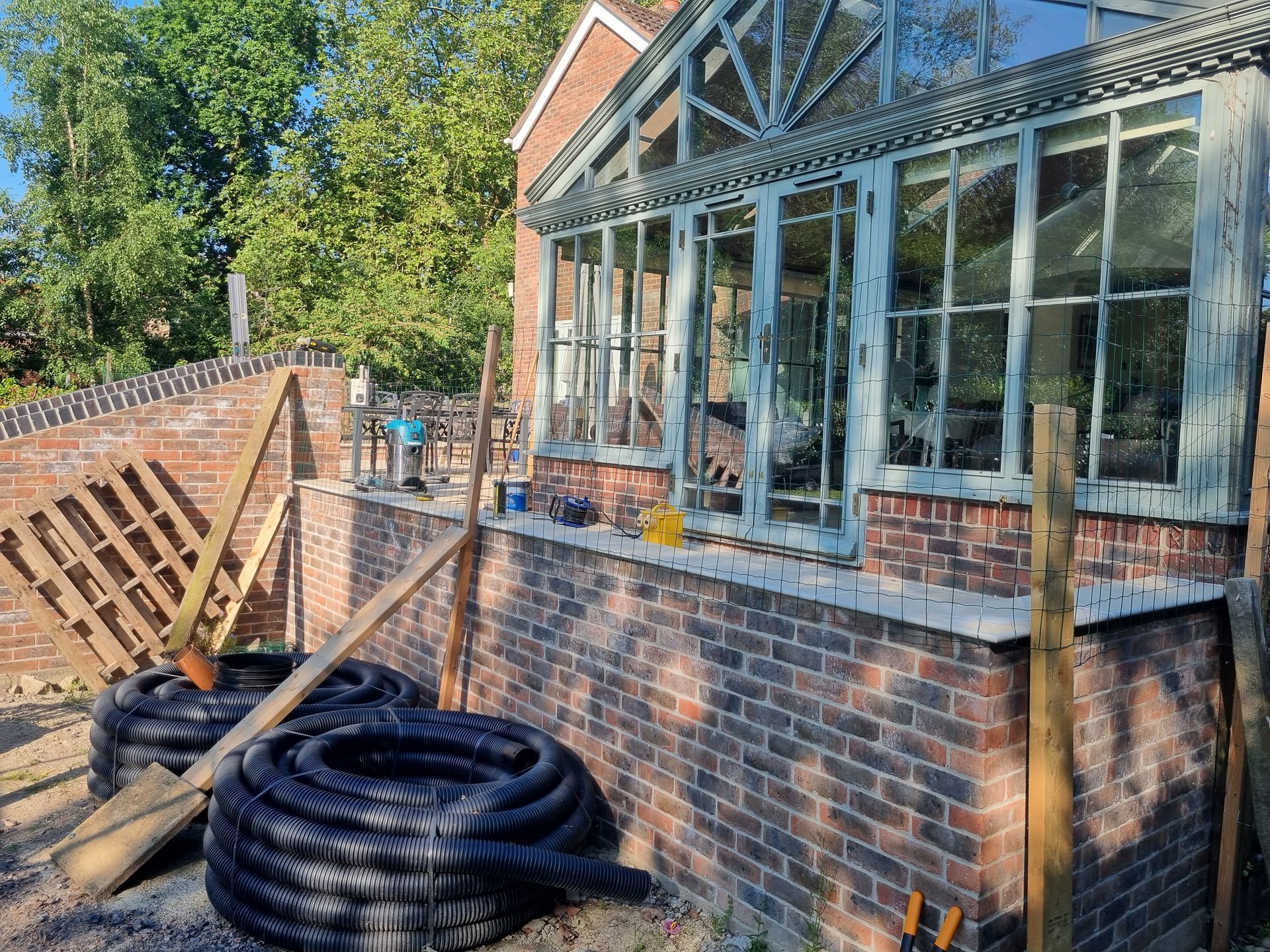Sunlit brick building with glass windows, under construction, with black coiled tubes and wooden supports.
