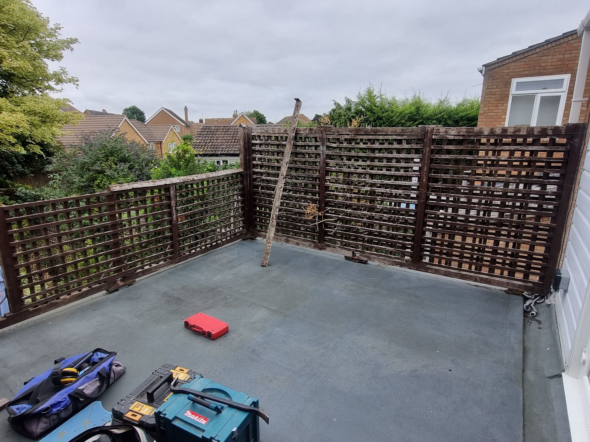 Dark wood lattice fence on gray flat roof; tools and a ladder present. Cloudy sky background.