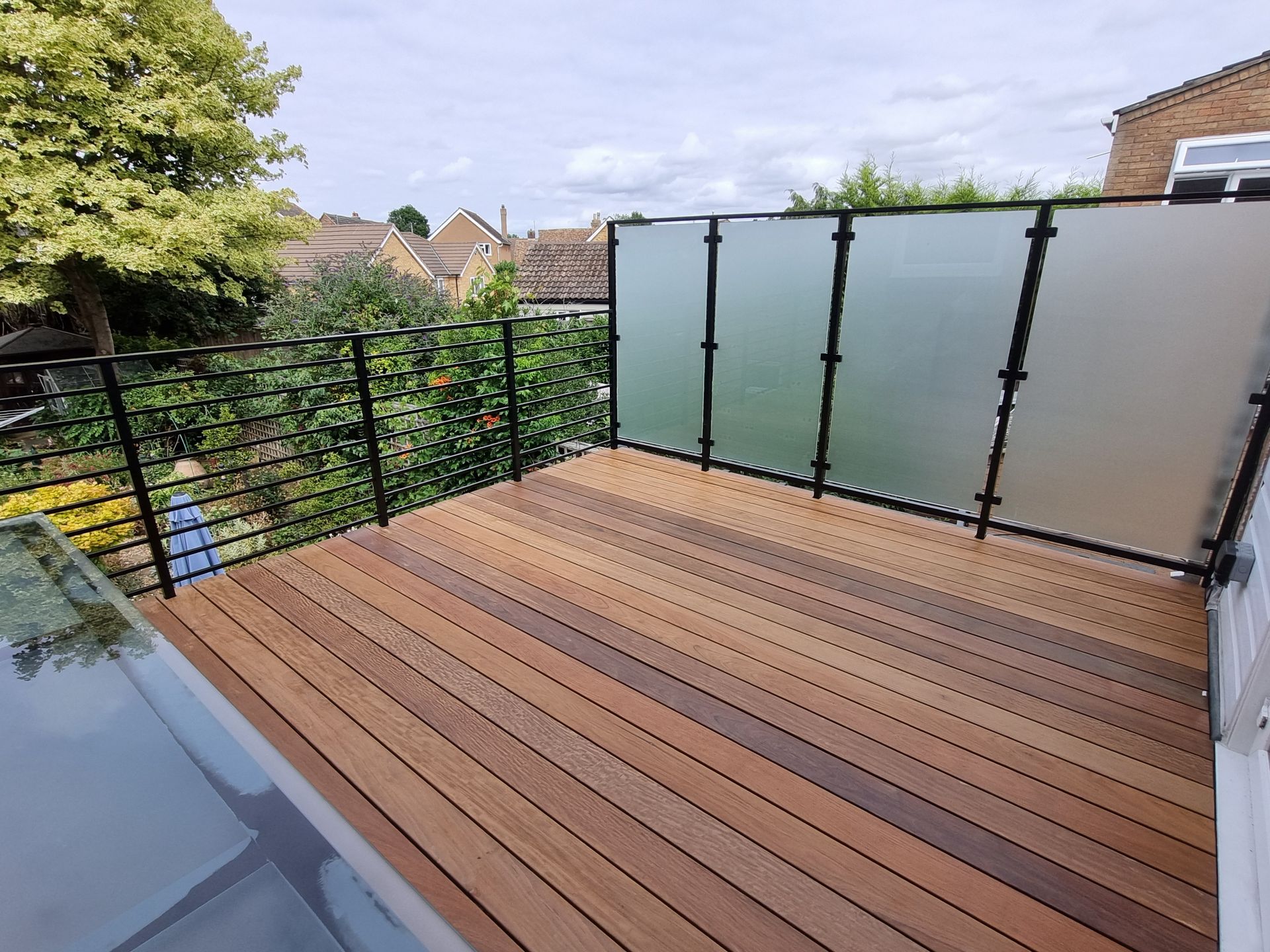Wooden deck with black railings and frosted glass panels, surrounded by greenery and houses.