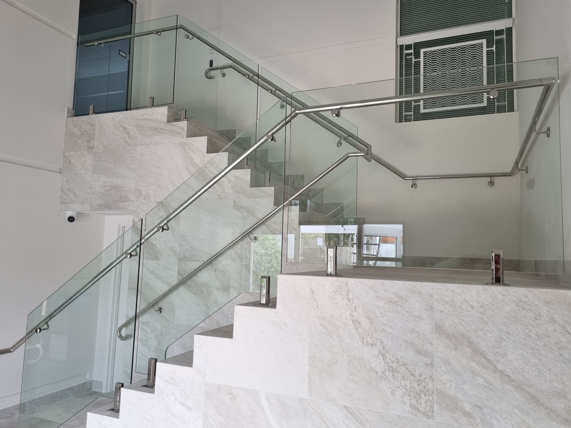 Staircase with marble steps, glass panels, and stainless steel handrails. White walls and natural light.