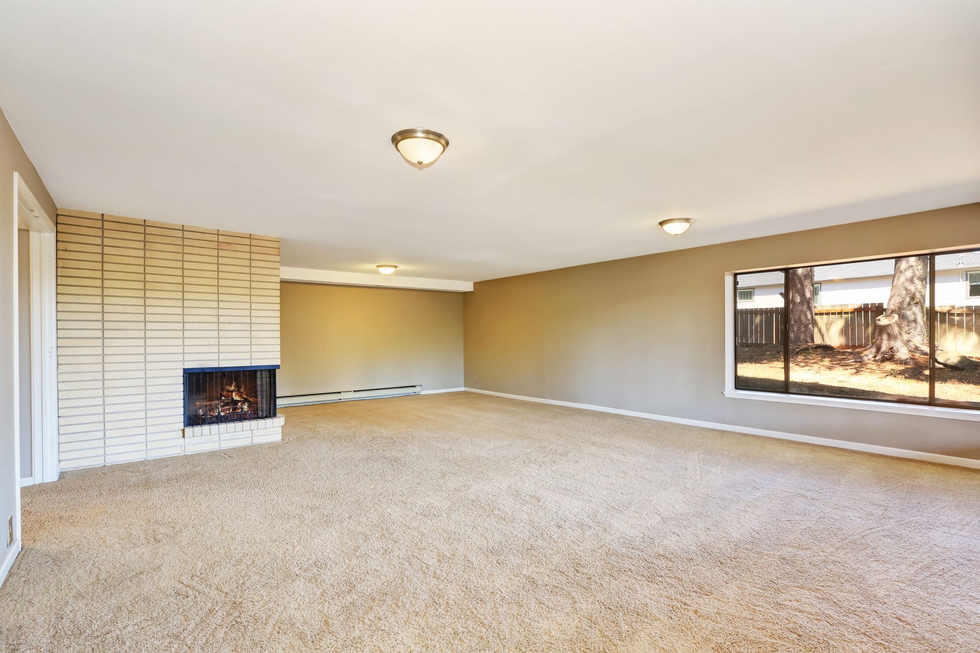 Empty beige living room with carpet, fireplace, and large window lighting the space