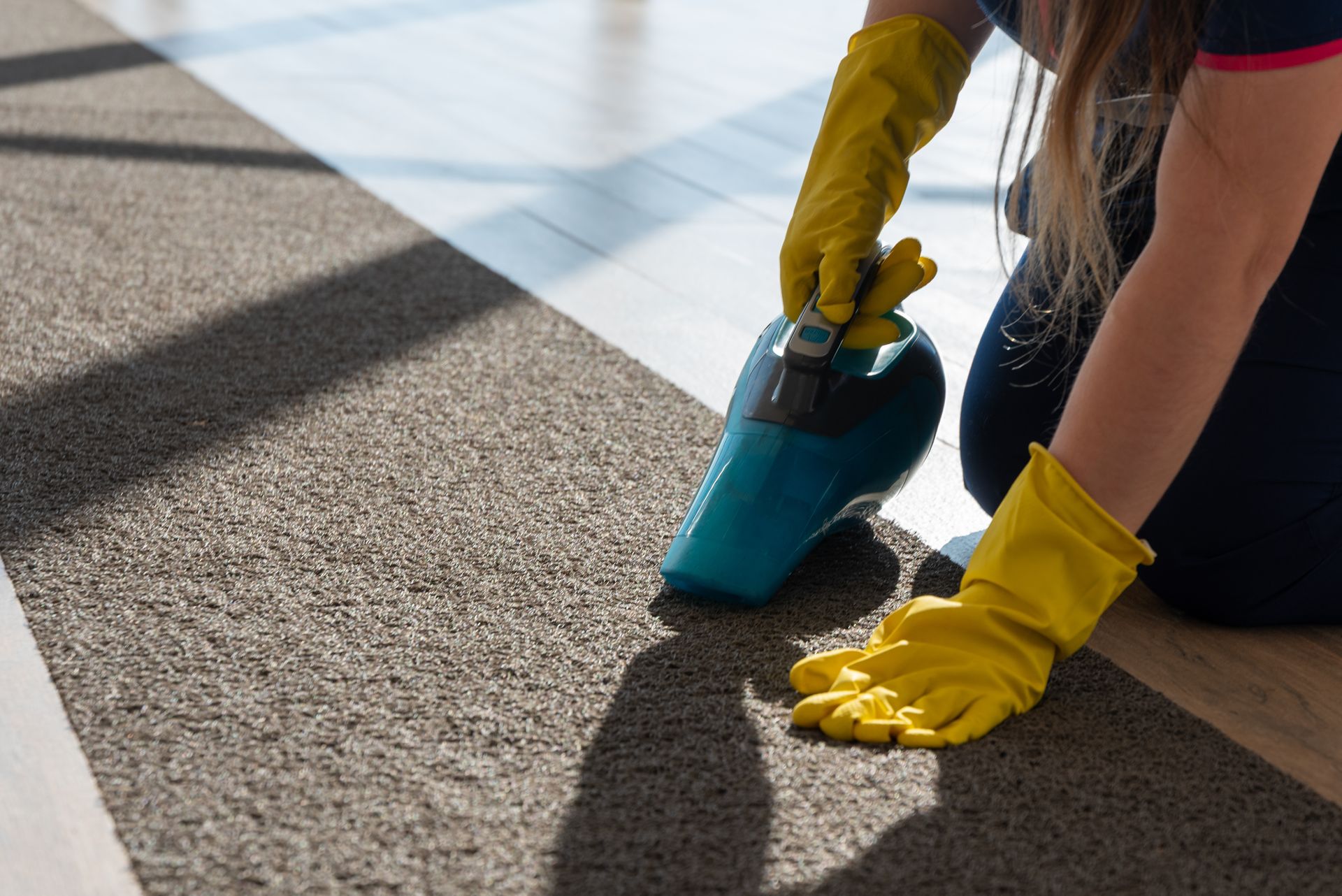 Person vacuuming a carpeted floor with a teal vacuum and yellow gloves in bright sunlight