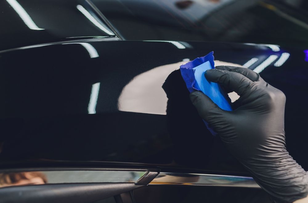 Person in black glove applying ceramic coating to a black car with a blue applicator.