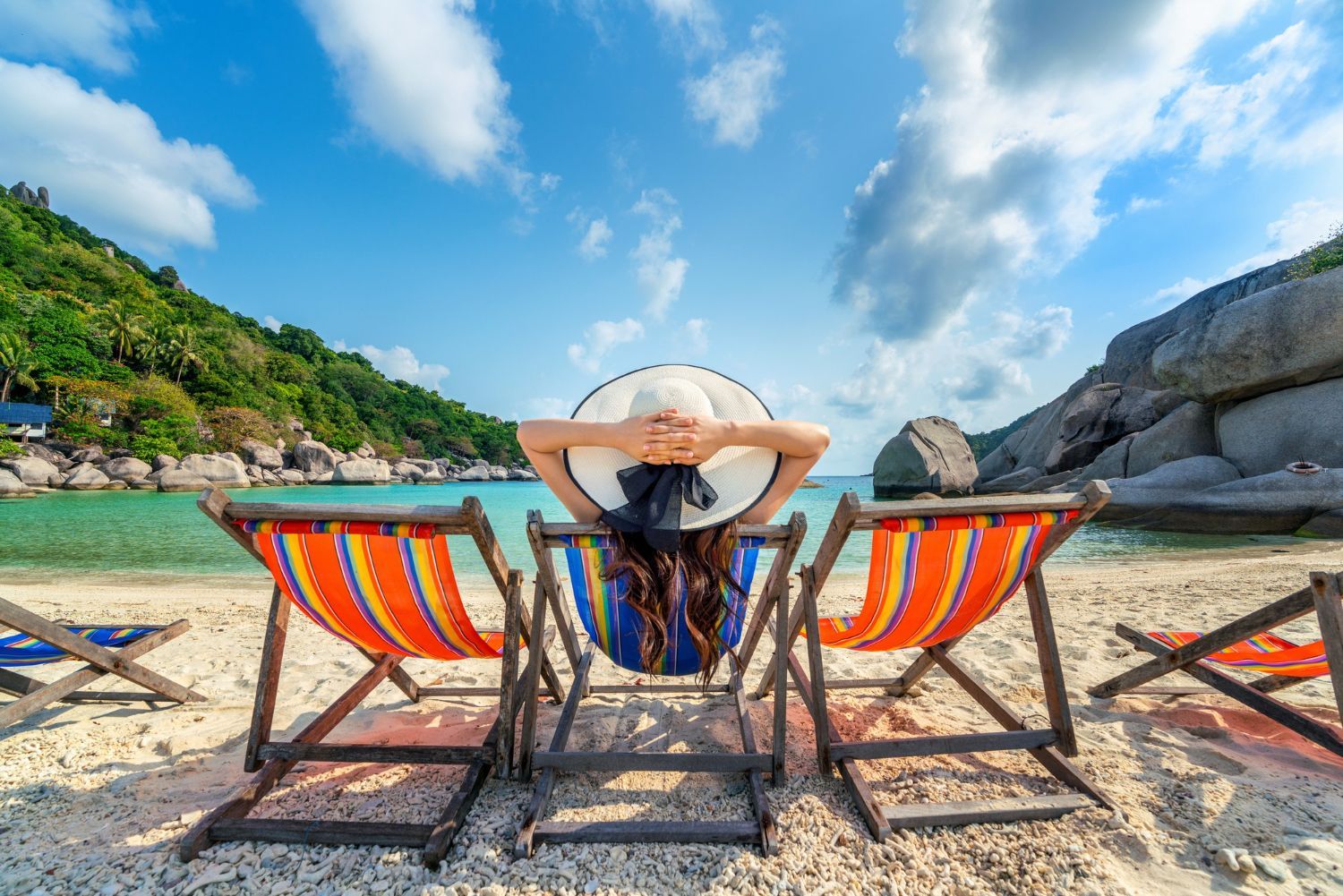 Woman in a sun hat relaxing in a beach chair, facing the ocean. Bright sky, white sand, and green hills.