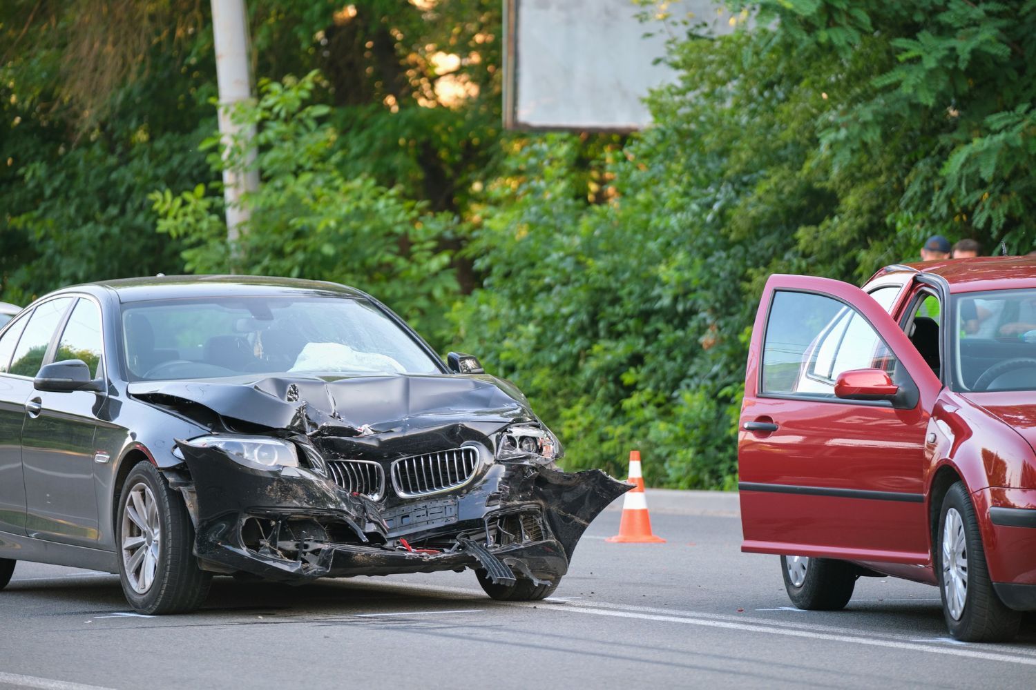 Damaged black car and red car on a road after a collision; orange traffic cone present.