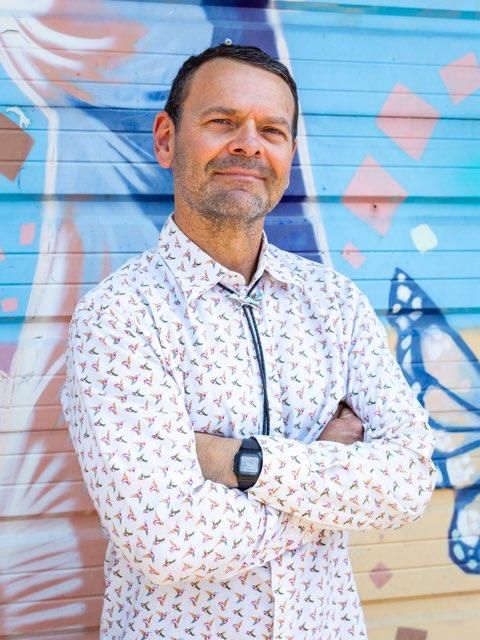Man with arms crossed, wearing a patterned shirt and bolo tie, standing in front of a colorful mural.