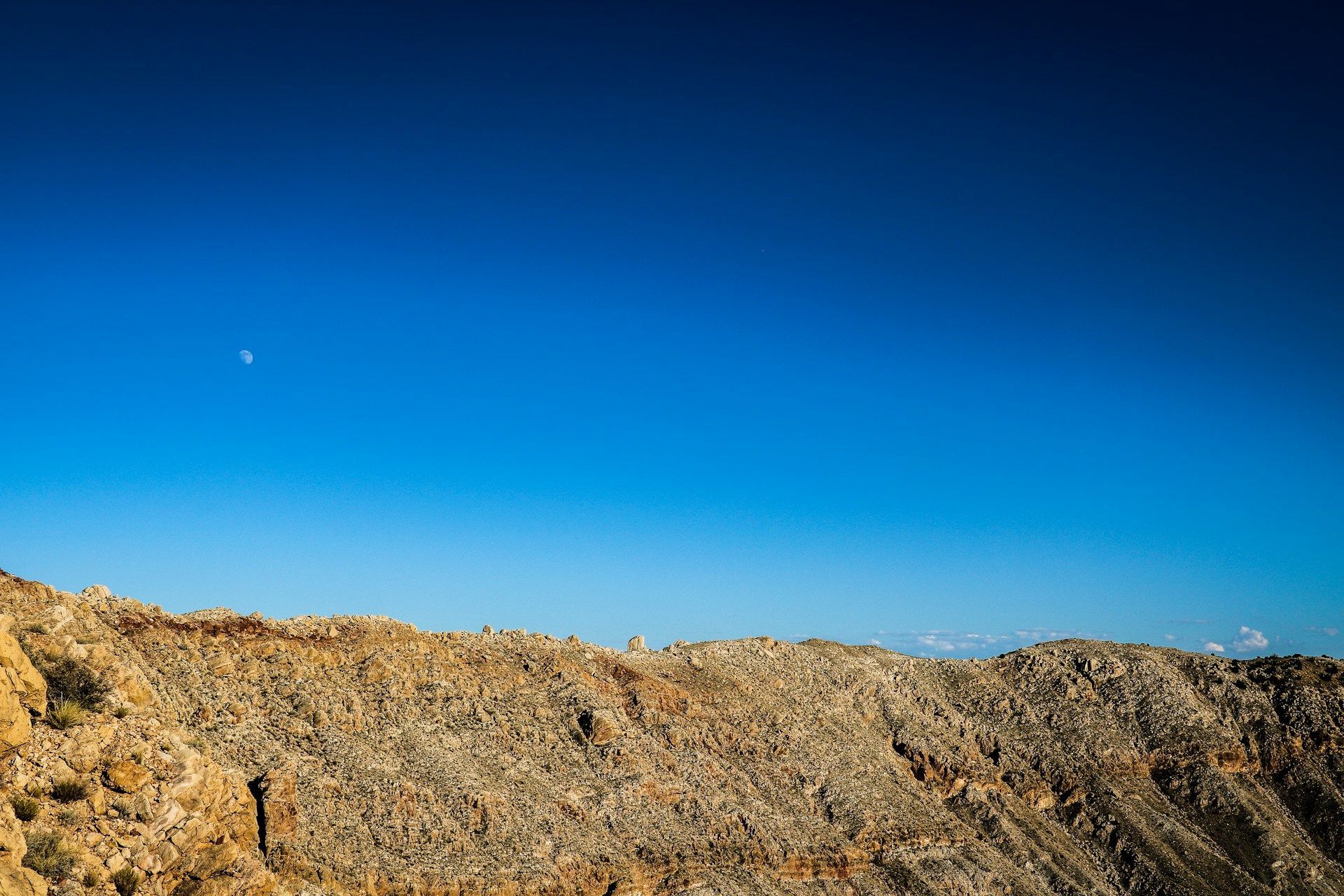 Rocky mountain ridge under a clear blue sky, with the moon and a bright planet visible.
