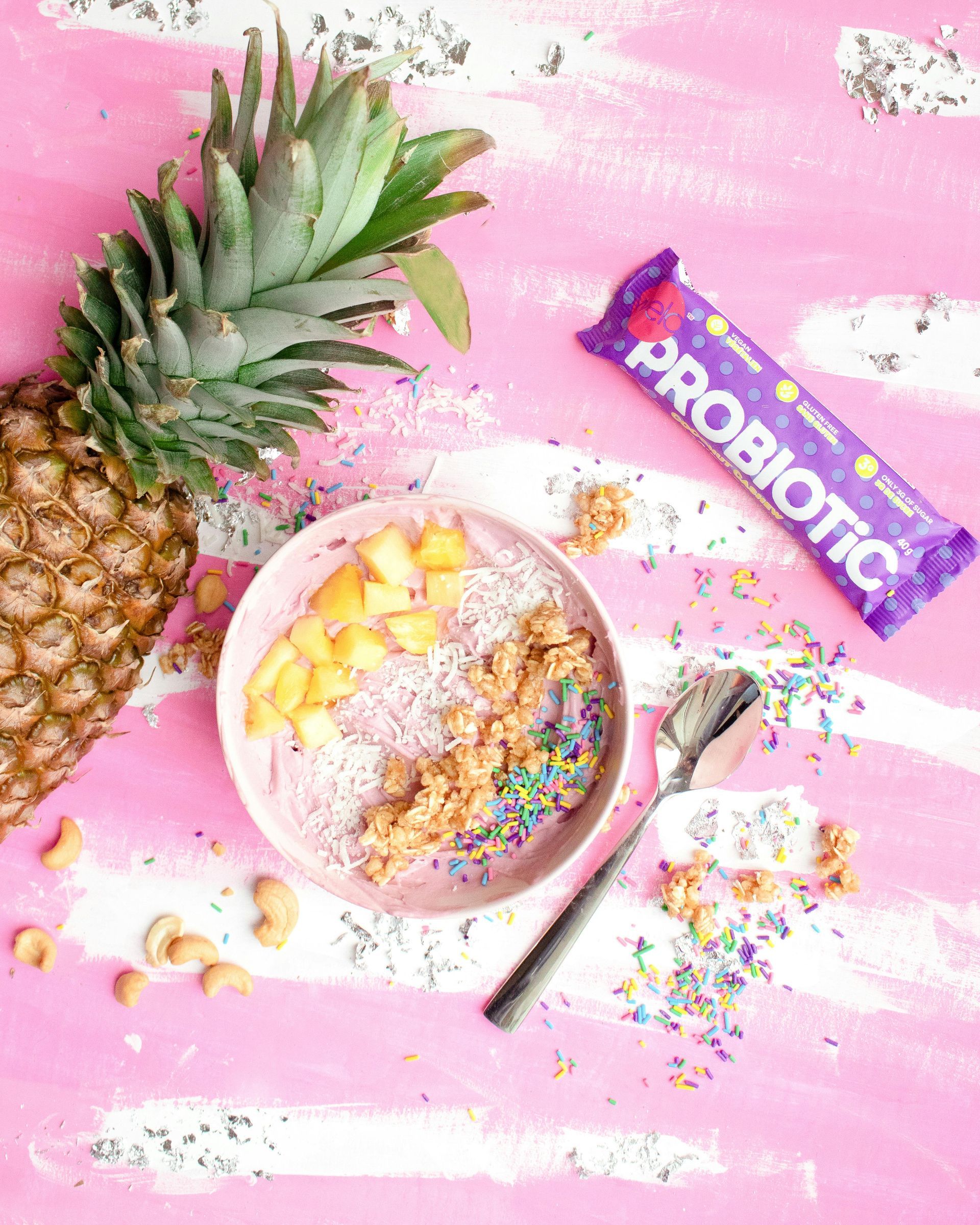 A pink smoothie bowl with pineapple, granola, and sprinkles, next to a pineapple and probiotic bar on pink.