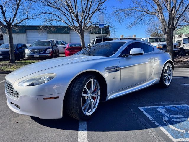 Silver sports coupe parked outdoors in a sunny lot with trees and other cars in the background