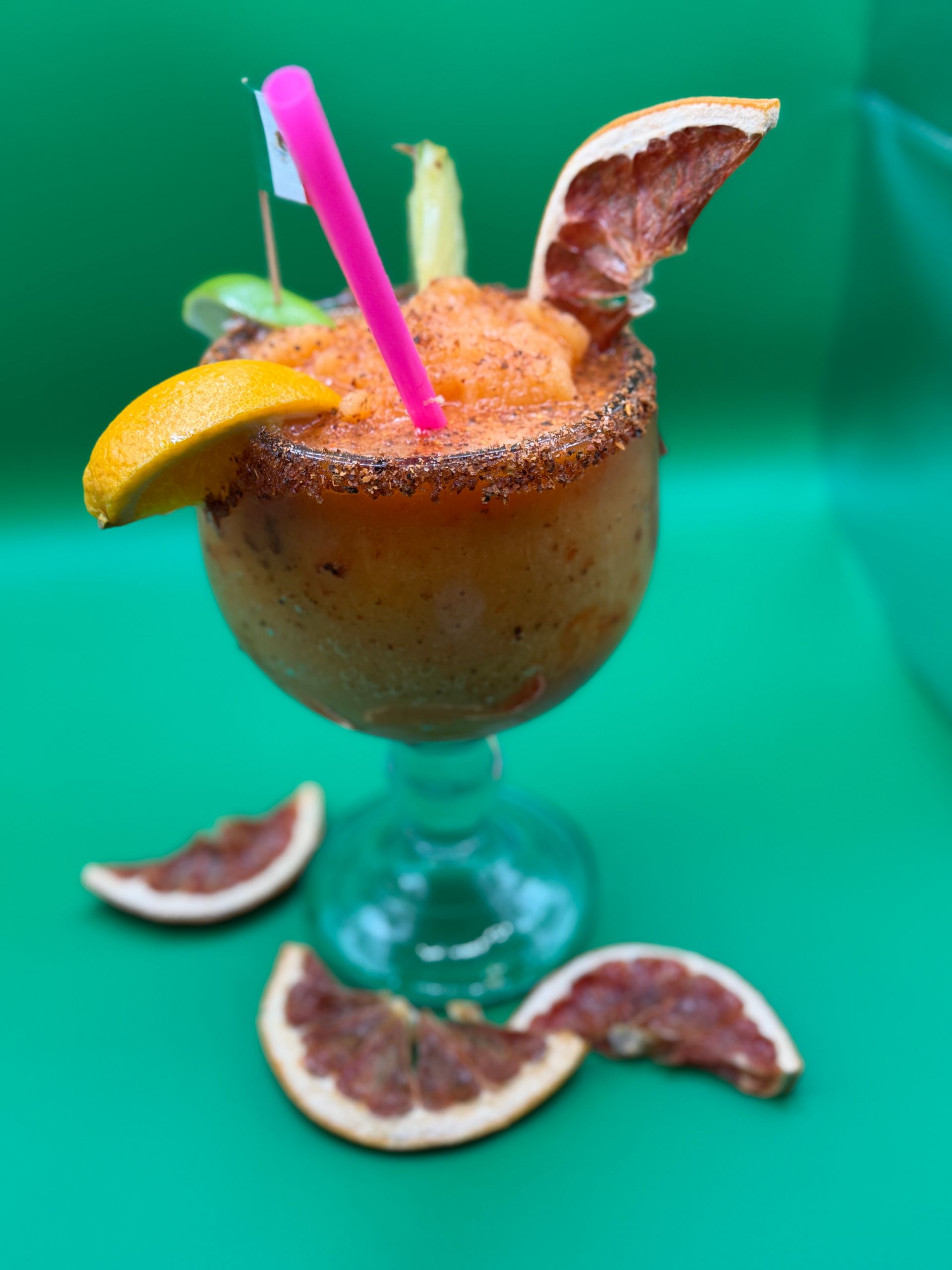 Tropical cocktail in a glass with pink straw and orange garnish, surrounded by passion fruit slices on green background