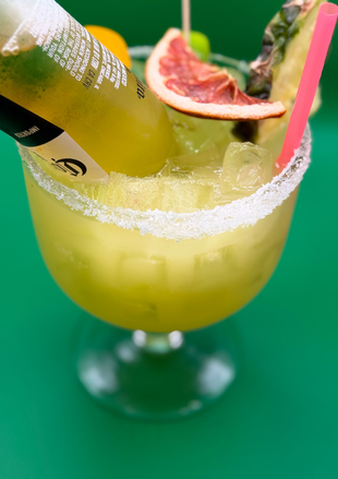 Yellow cocktail with a salted rim, pink straw, and grapefruit garnish on a green table.