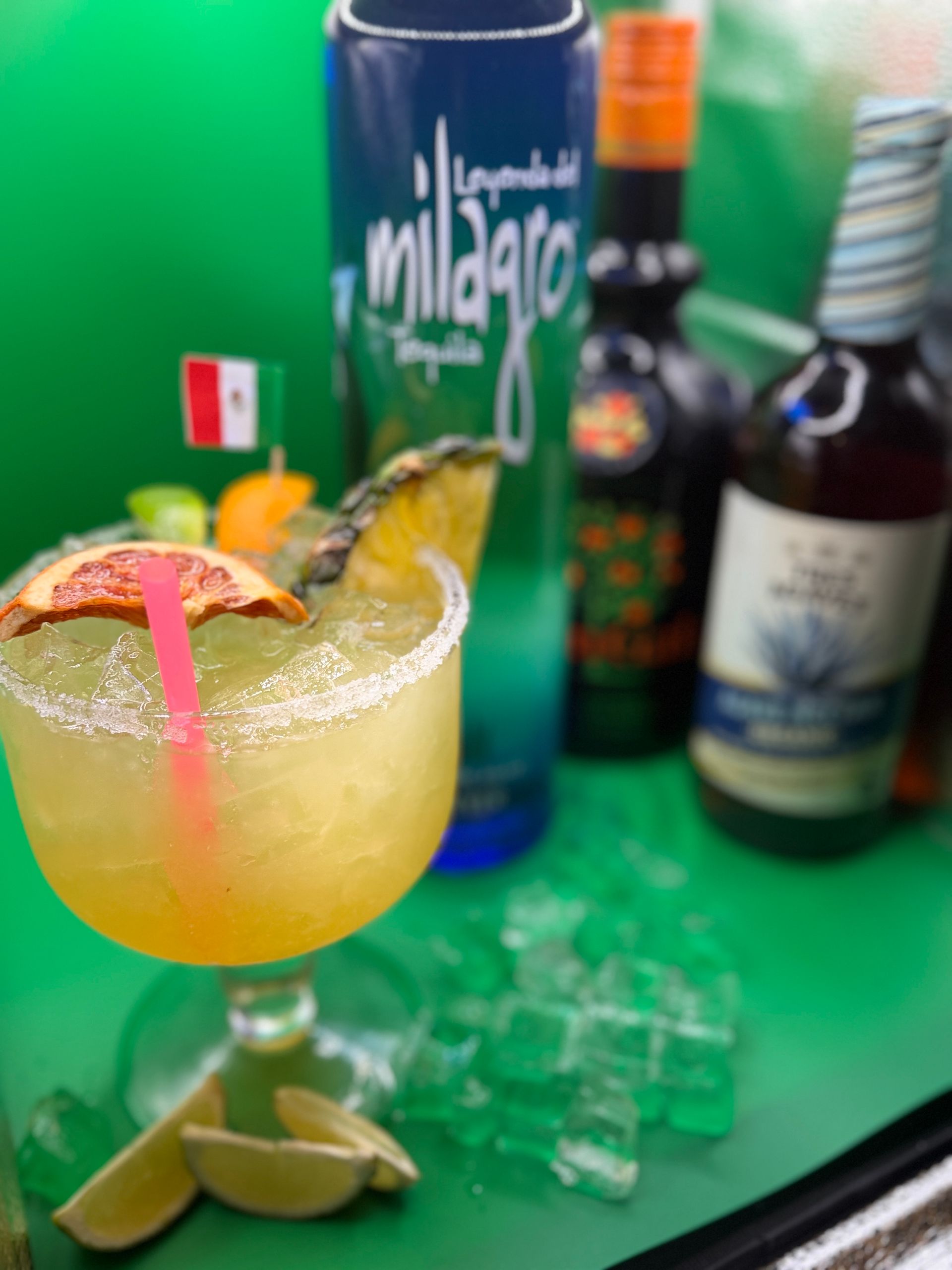Cocktail with lime and red straw on a green table, with liquor bottles and ice in the background