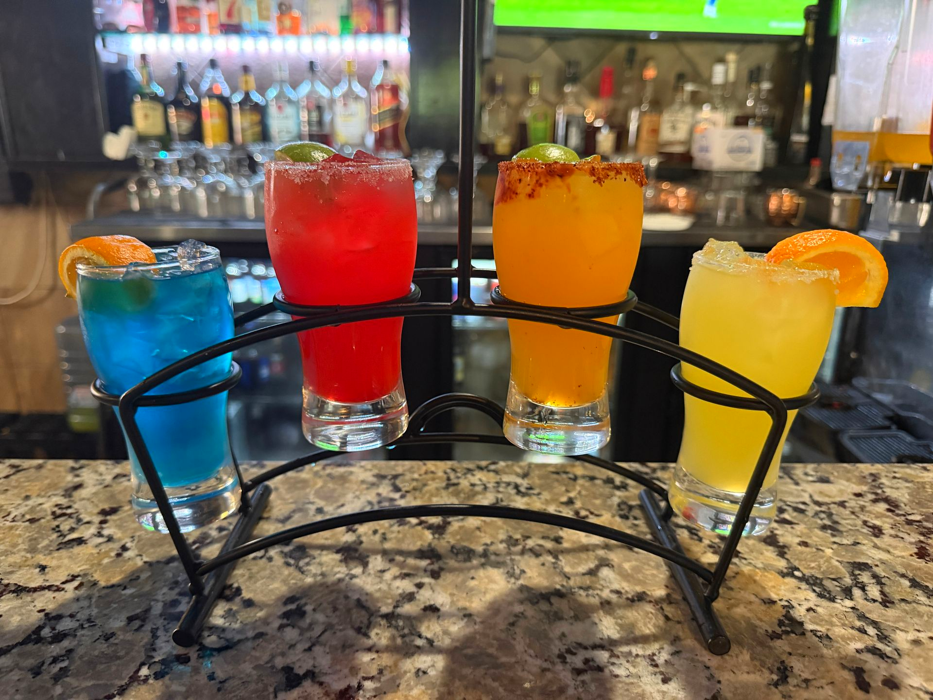Four colorful cocktails with fruit garnishes on a granite bar, arranged in a curved black rack.