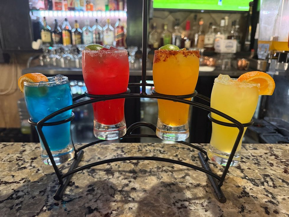 Four colorful cocktails in a black metal holder on a bar counter, with bottles blurred in the background
