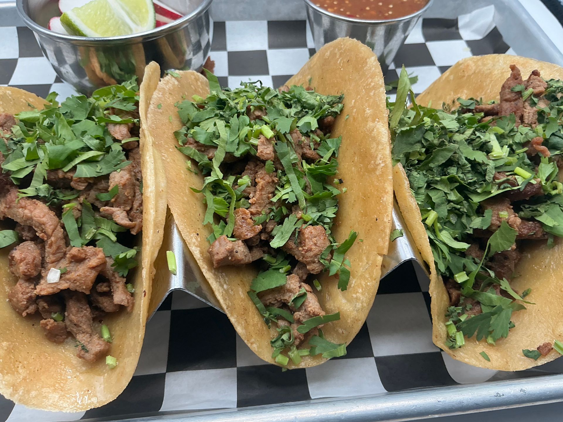 Three beef tacos topped with cilantro on a checkered tray, with lime wedges and salsa in the background