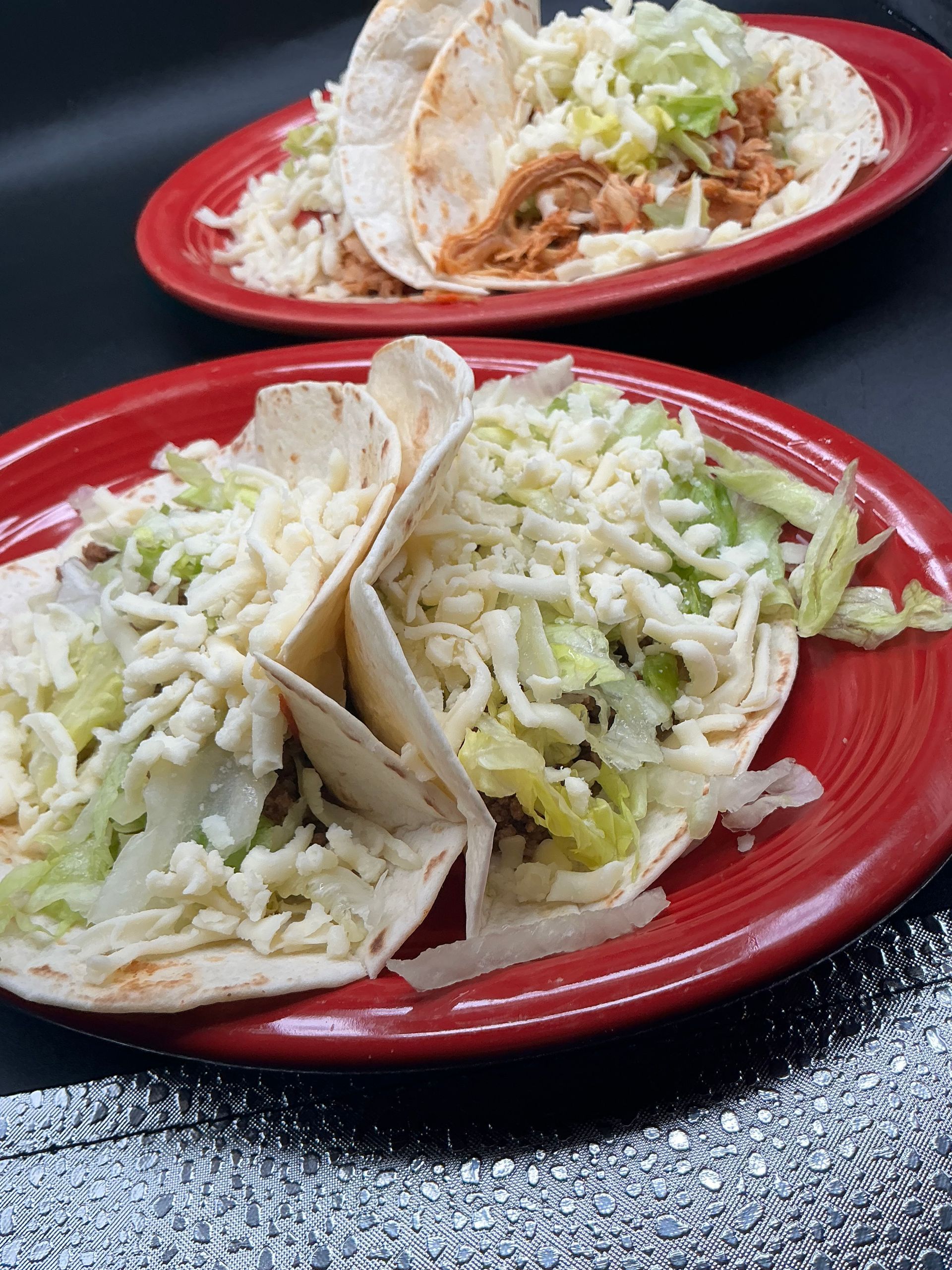Two red plates with chicken tacos topped with shredded lettuce and cheese on a dark table