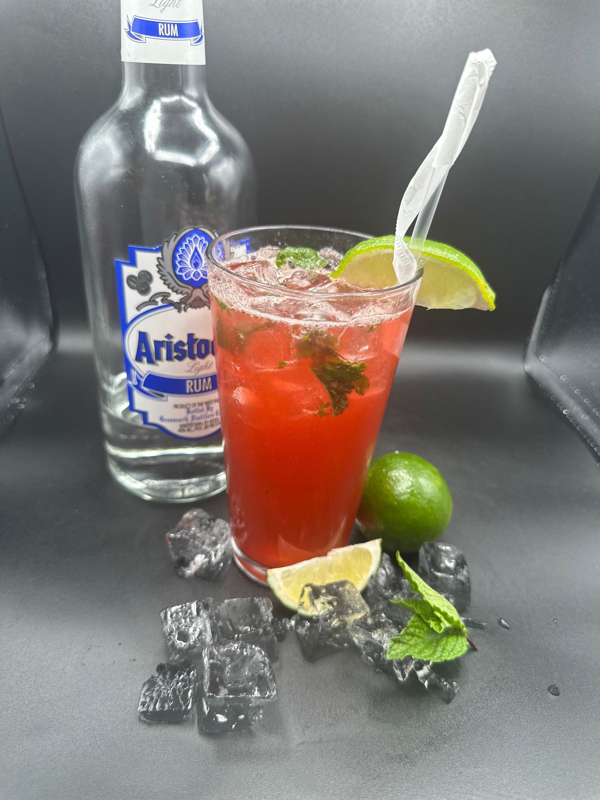 Red strawberry drink with lime and straw beside a bottle, ice, and lime wedges on a dark surface