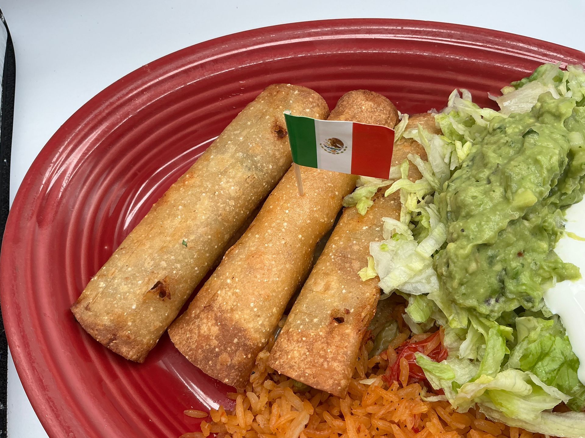 Three taquitos on a red plate with shredded lettuce, guacamole, and a small Mexican flag garnish