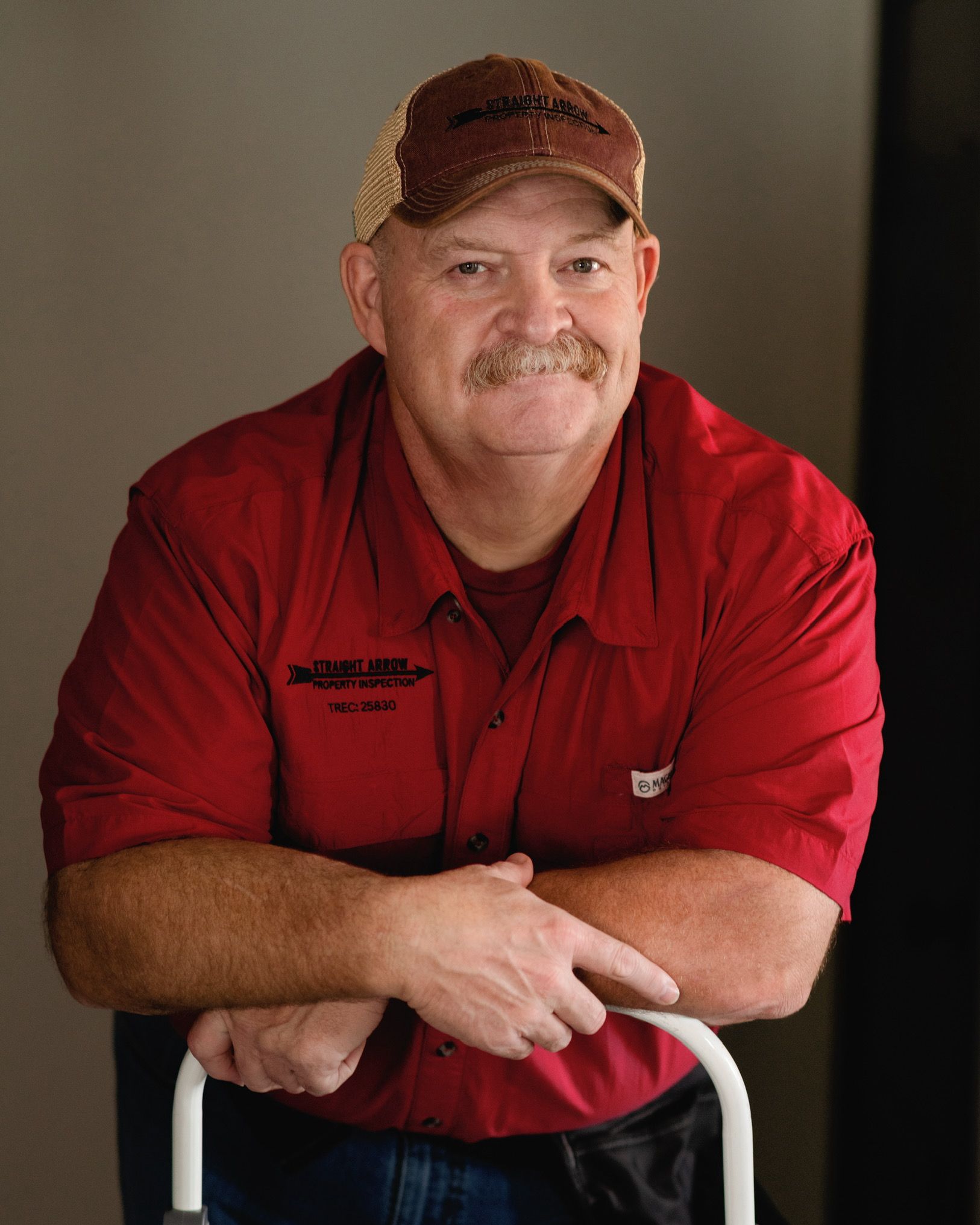 Man in red shirt and cap smiles, leaning on a white object; brown cap, mustache.