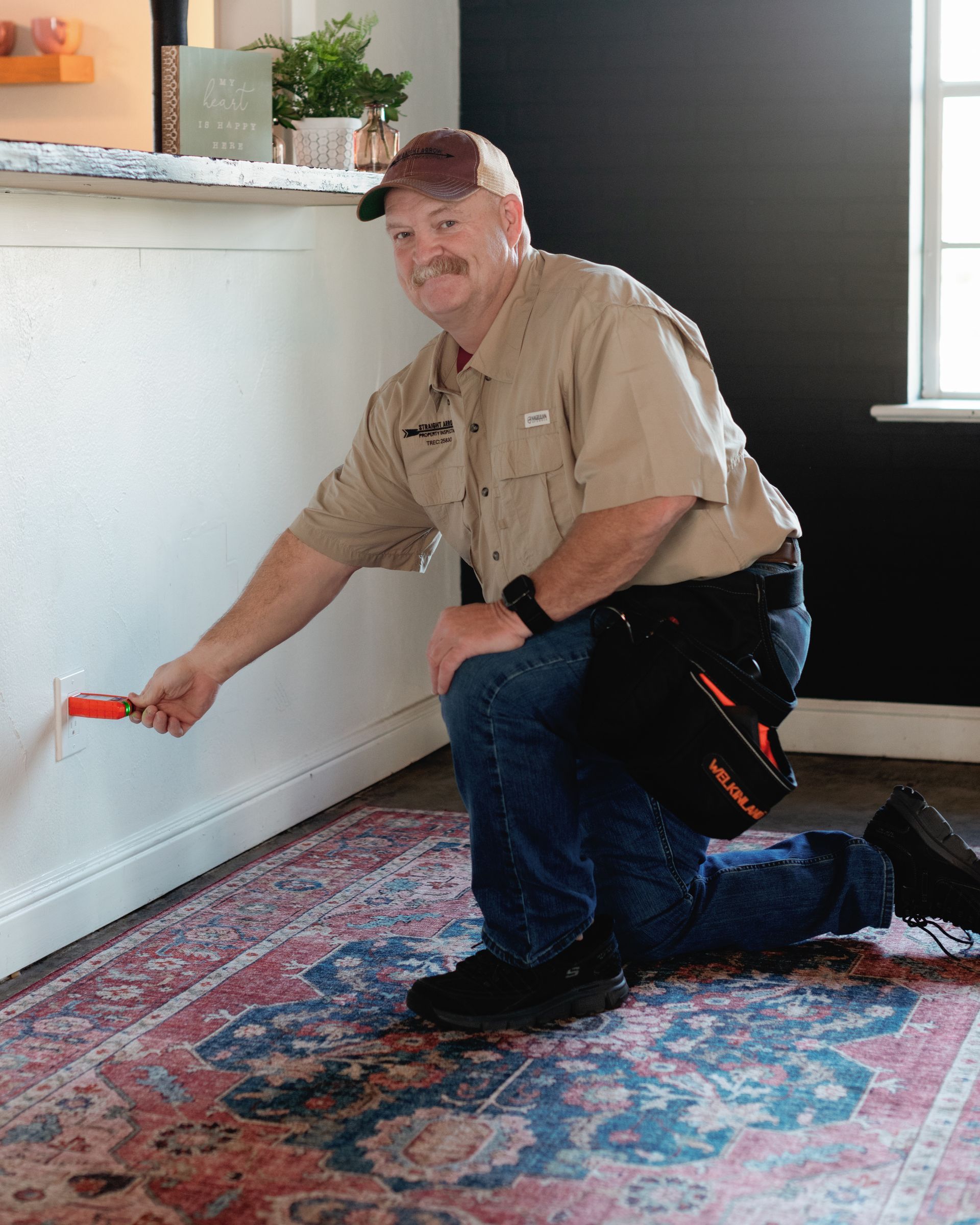 Man kneeling, testing electrical outlet with a tool. Wearing tan shirt, jeans, and hat. In a home interior.
