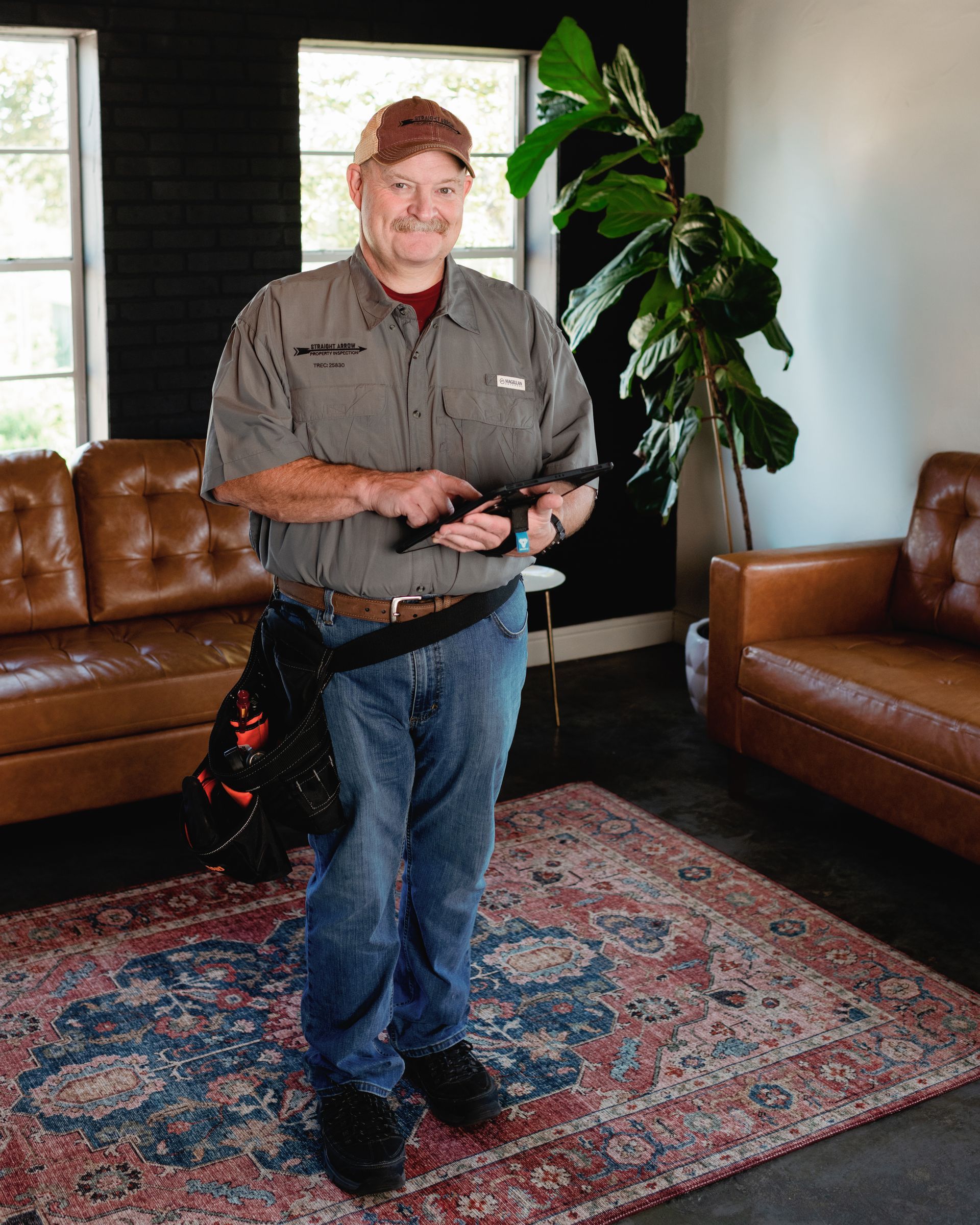 Man in work uniform holding tools, standing on a rug. Brown couches, indoor plant in background.