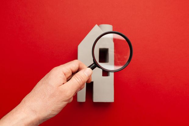 Hand holding magnifying glass over a white house cutout against a red background.