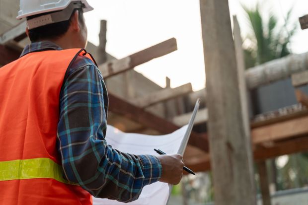 Construction worker in safety vest and helmet examines blueprints at a construction site.