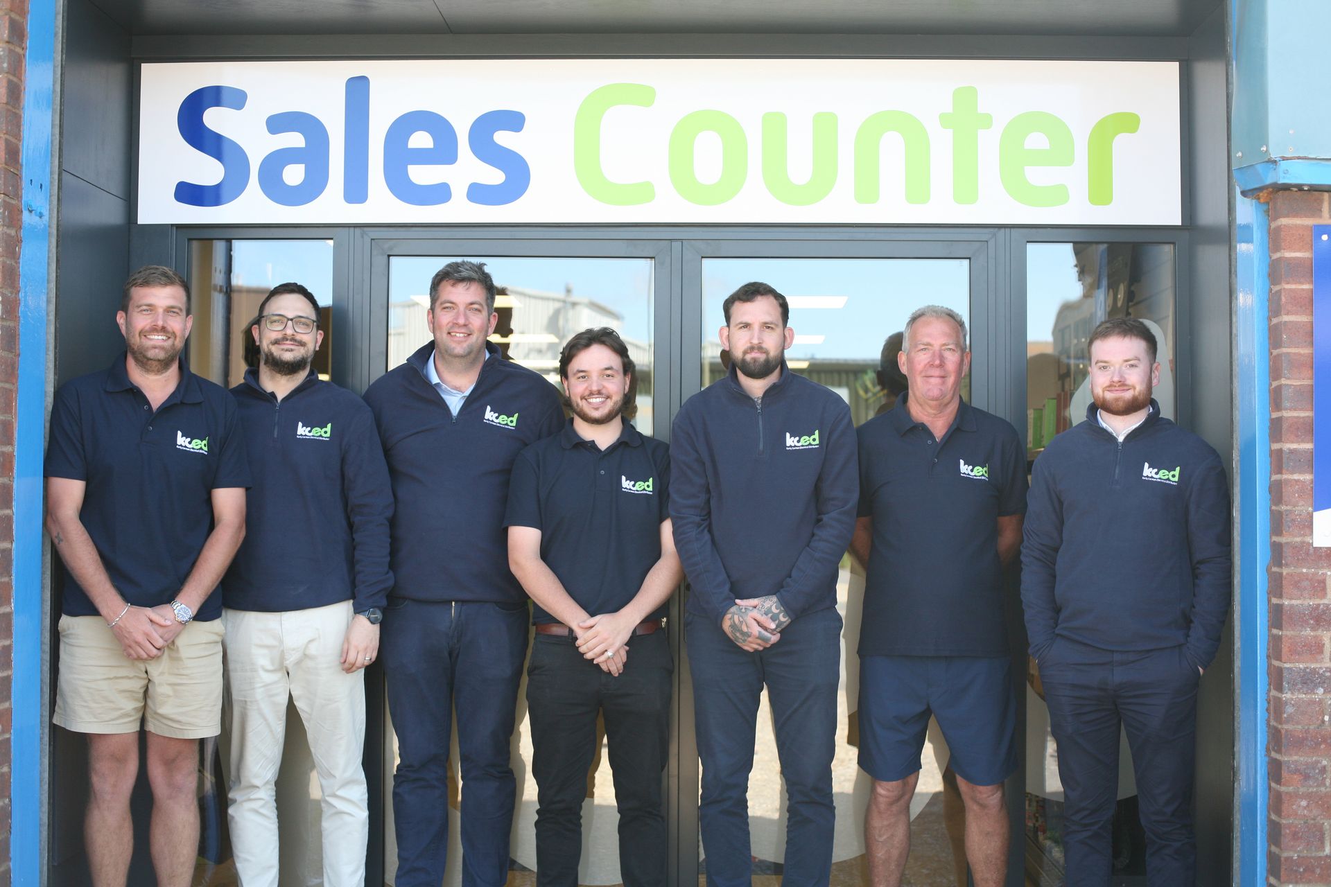Seven employees standing in front of a storefront sign reading “Sales Counter”