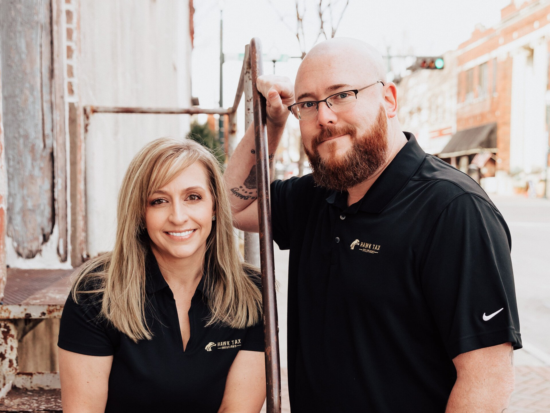 Two people in black shirts standing by a brick wall, smiling outdoors in daylight.