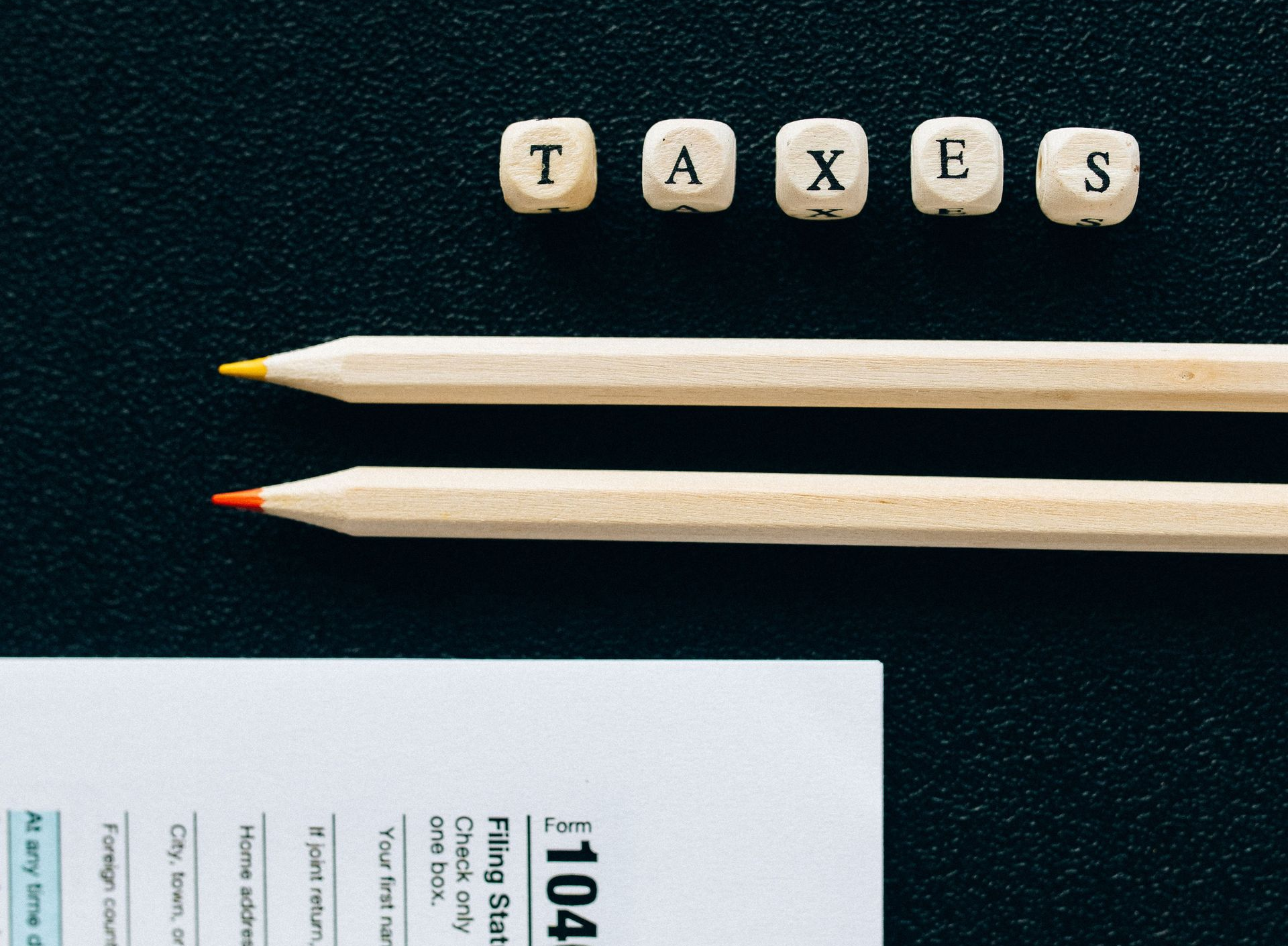 Wooden letter tiles spelling “TAXES” above two pencils on a dark desk, with paper at bottom left