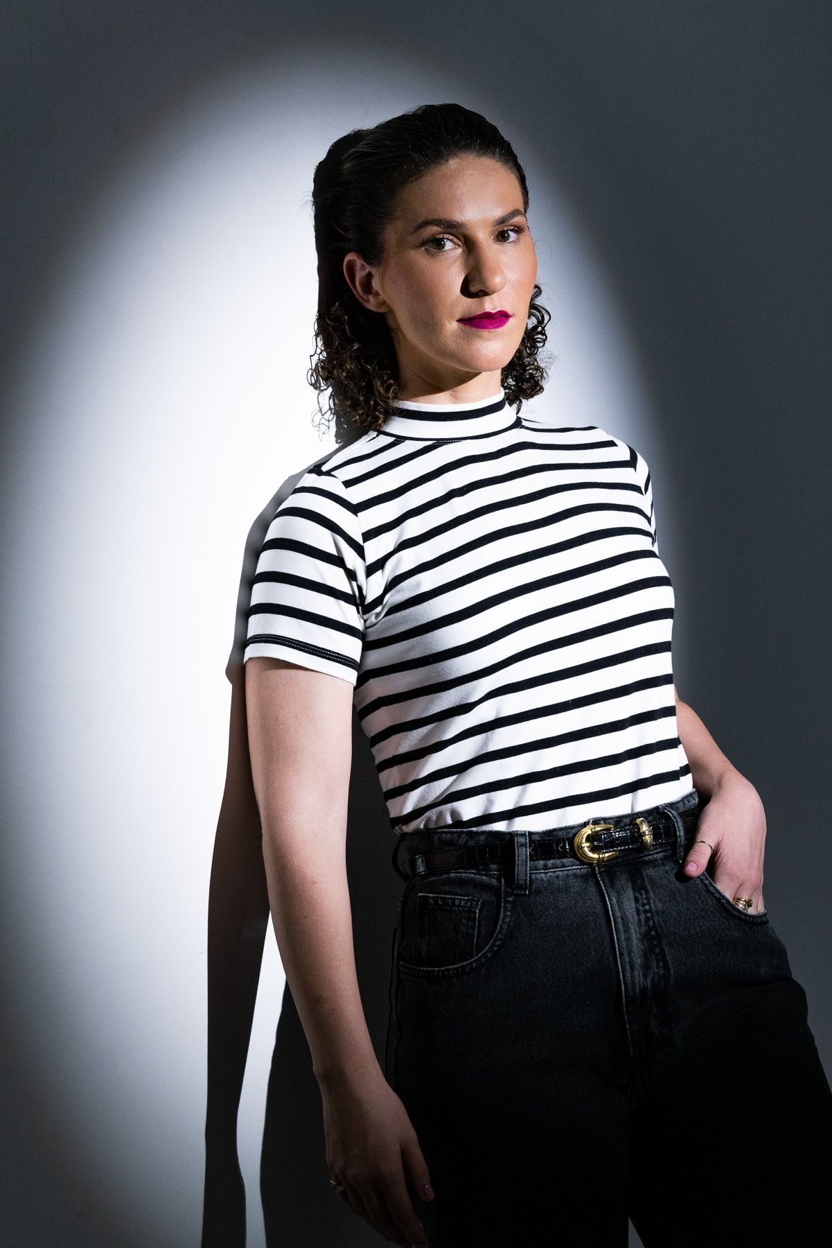 Nayara Falconi de camisa listrada e jeans está parada em frente a uma parede.