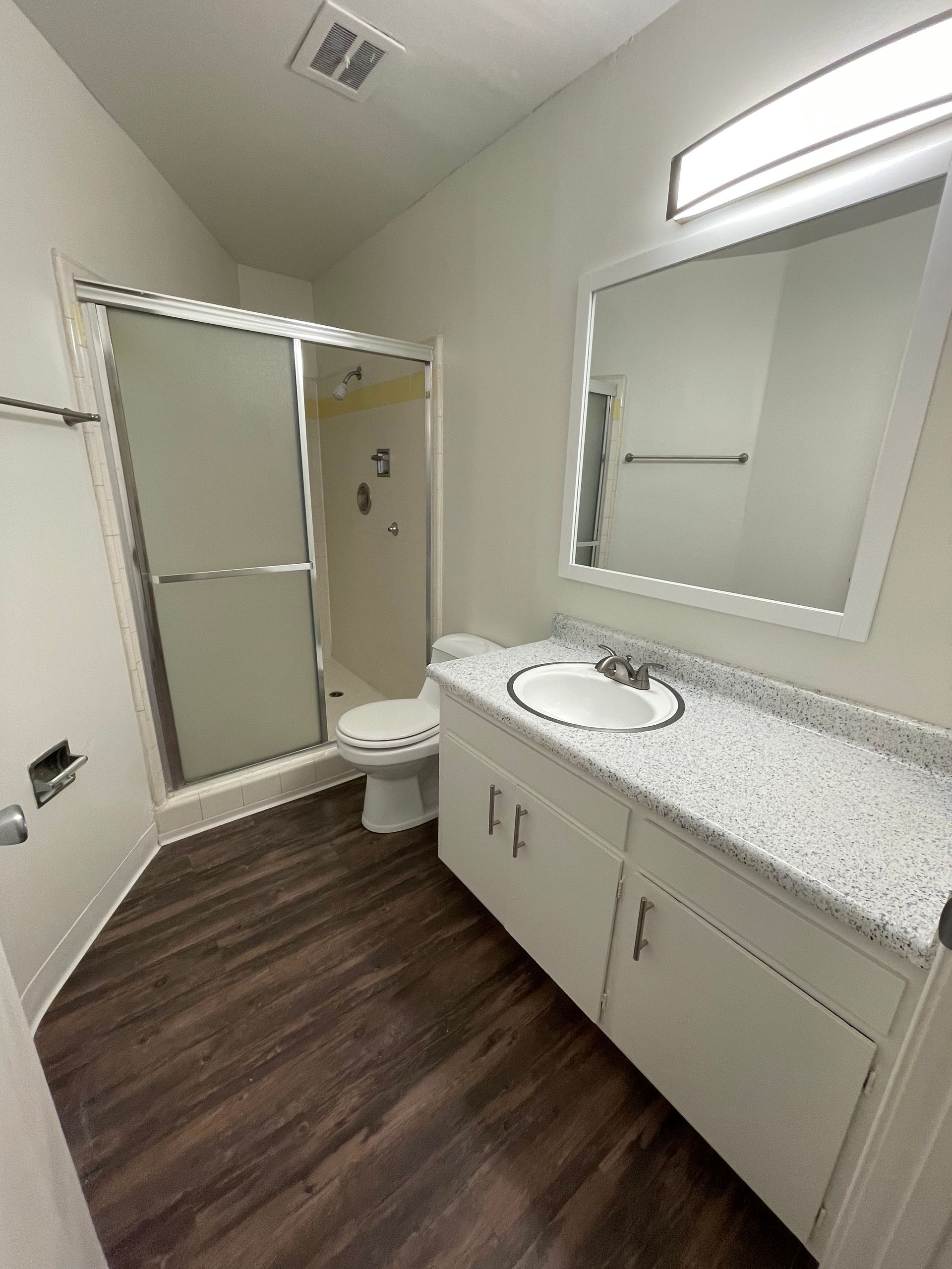 Small bathroom with white vanity, mirror, toilet, and shower; dark wood-look floor and speckled countertop.