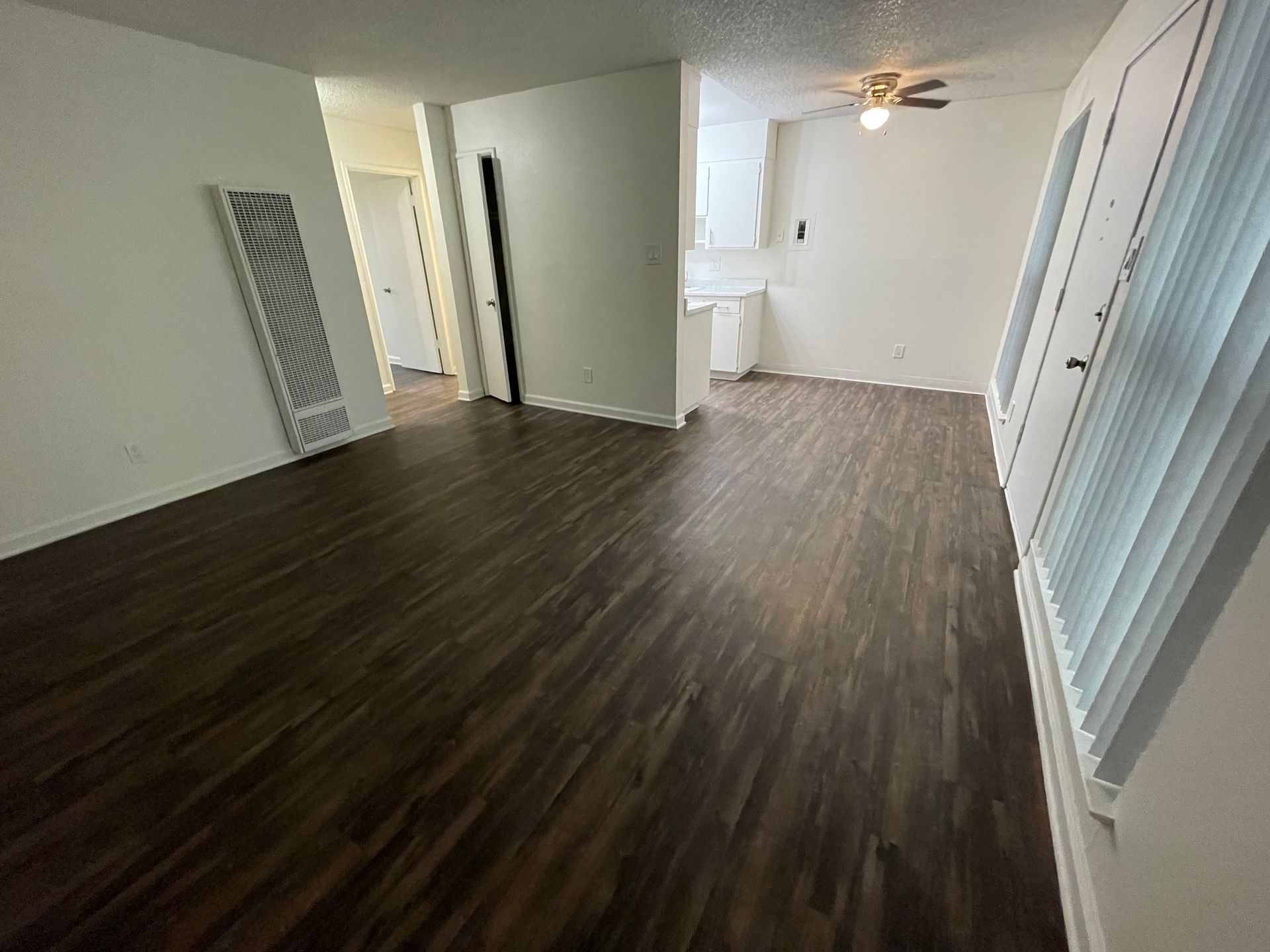 Empty living room with dark wood floors, white walls, ceiling fan, and blinds by the window.