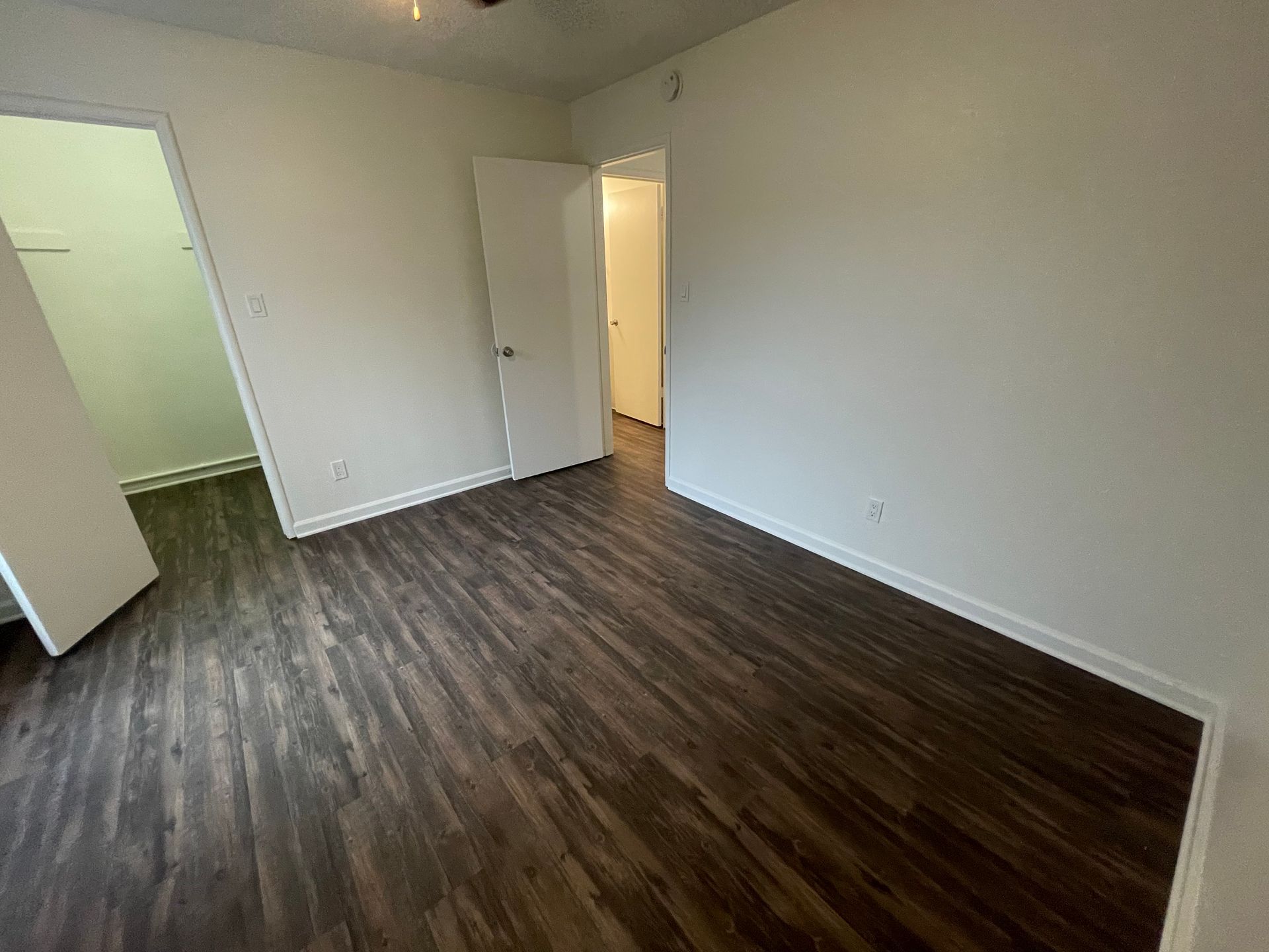 Empty room with dark wood flooring, white walls, and an open doorway to a lit hallway.