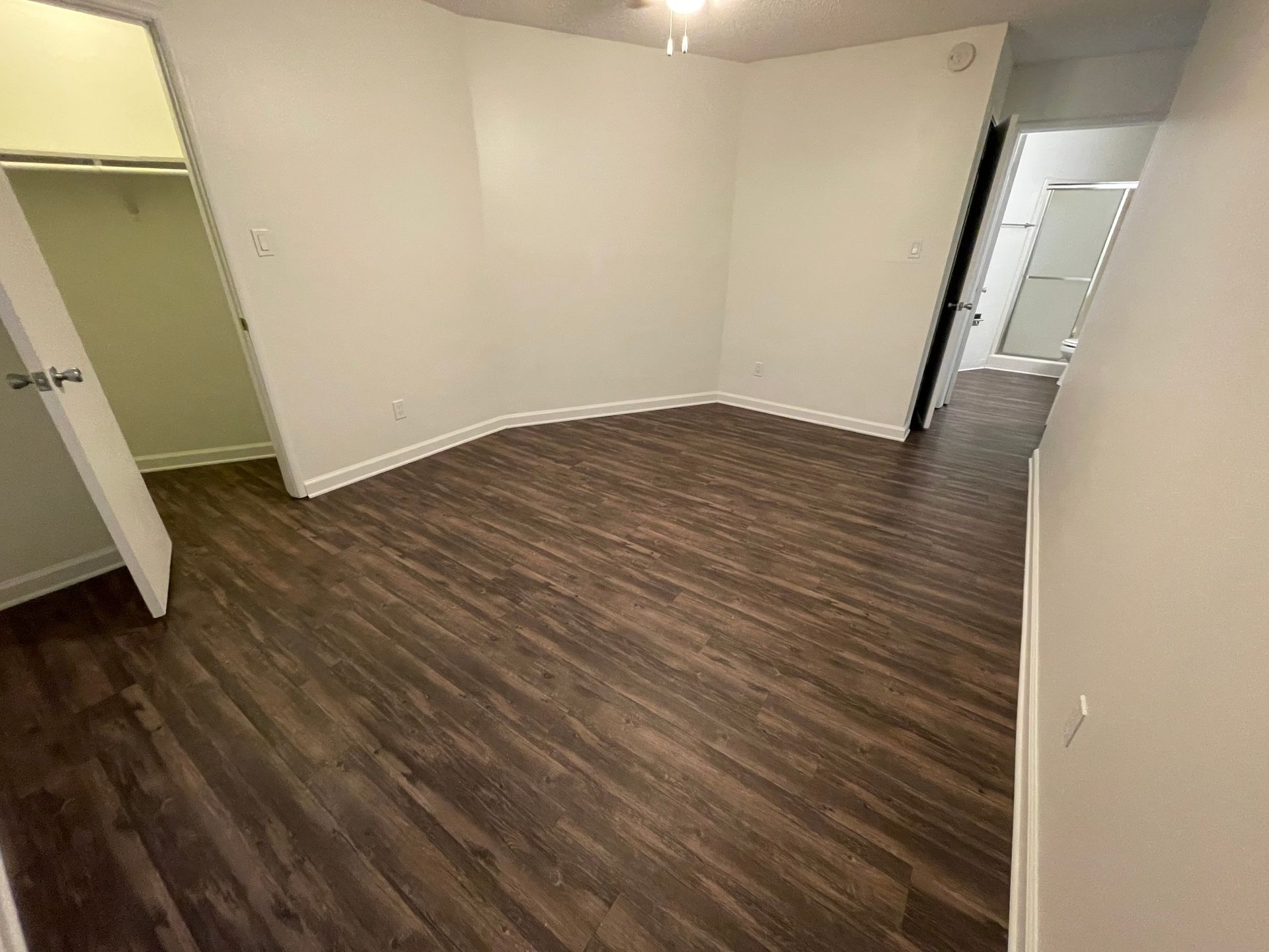 Empty room with brown wood-look flooring, beige walls, and a doorway to a hall on the right.