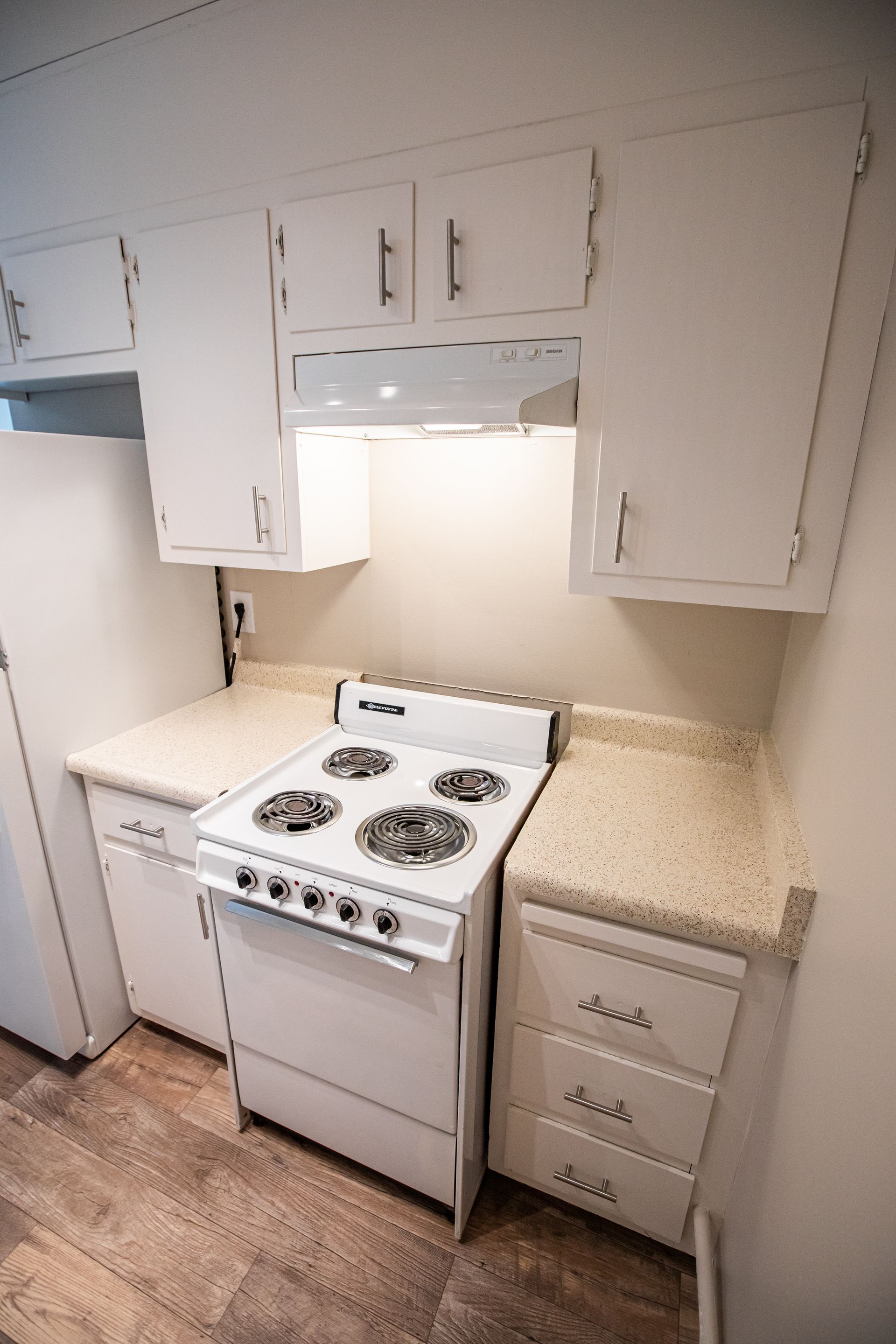 Small vintage kitchen with white cabinets, stove, refrigerator, and speckled countertops under a range hood.